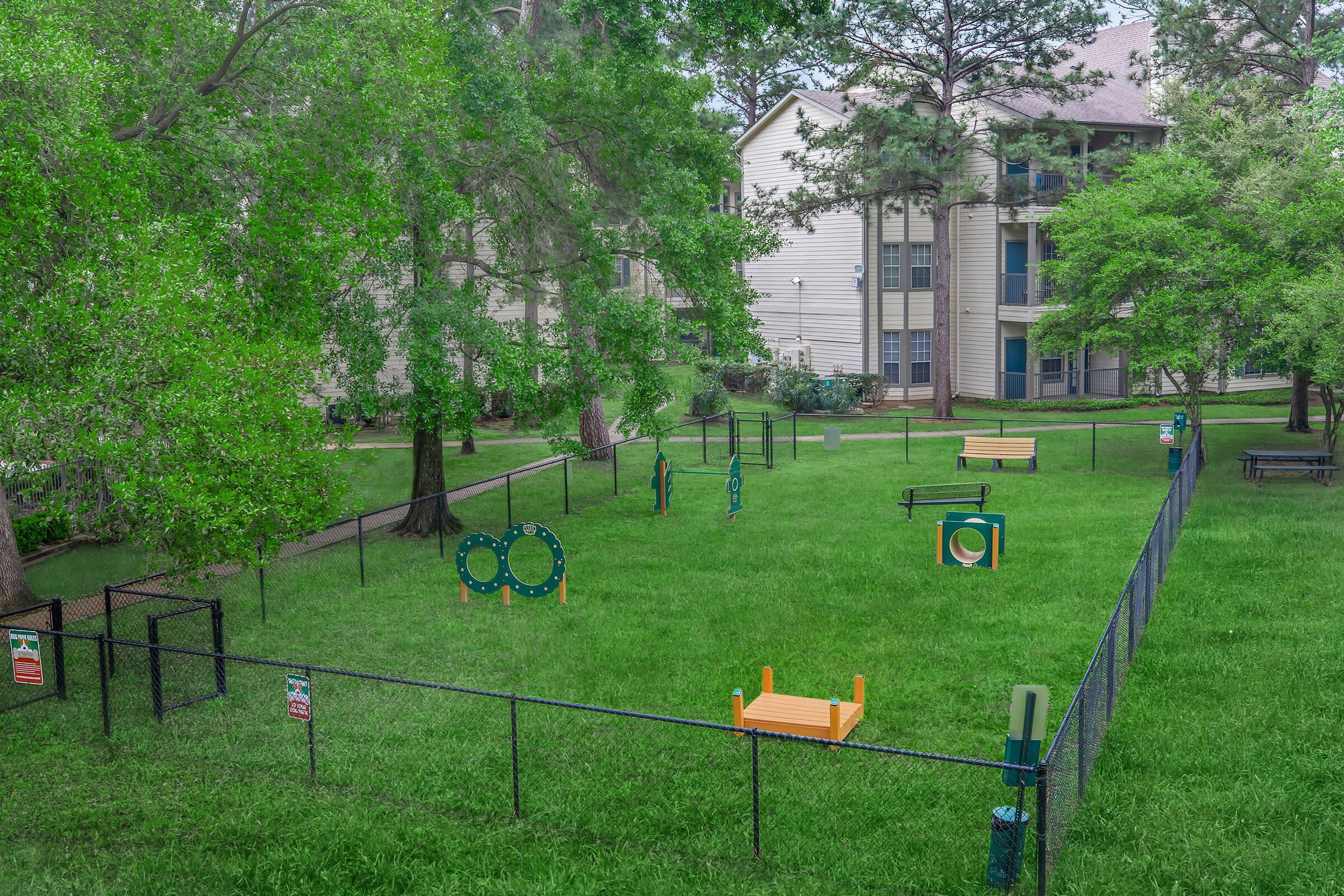 A fenced grassy area featuring various play structures and benches, surrounded by trees and apartment buildings. The space is designed for recreational activities, with equipment positioned throughout the yard. The greenery enhances the inviting atmosphere of the area.