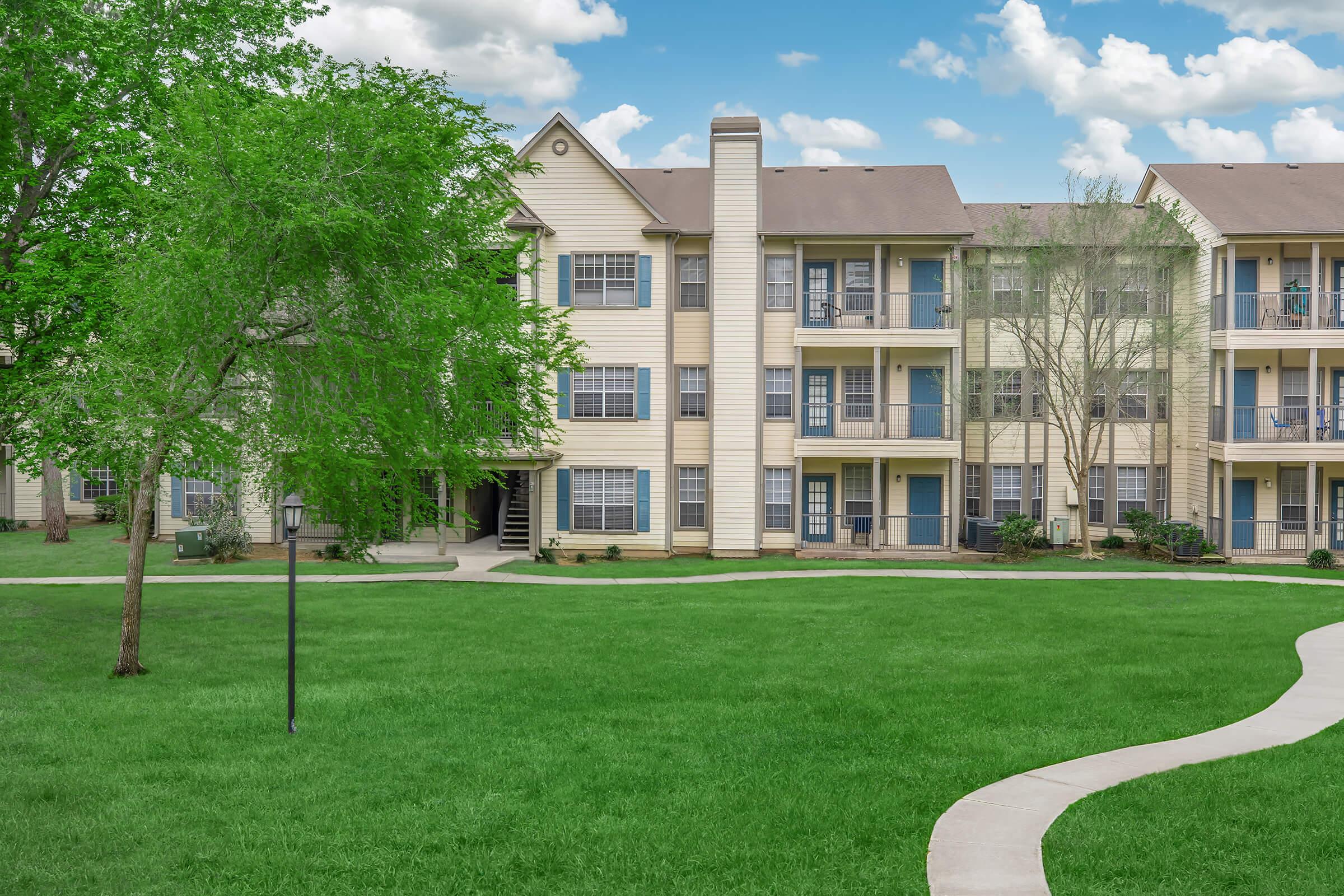A well-maintained apartment complex featuring several multi-story buildings with beige siding. Lush green grass surrounds the buildings, and a paved walkway leads through the landscape. There are trees providing shade, and the sky is partly cloudy, creating a pleasant atmosphere.