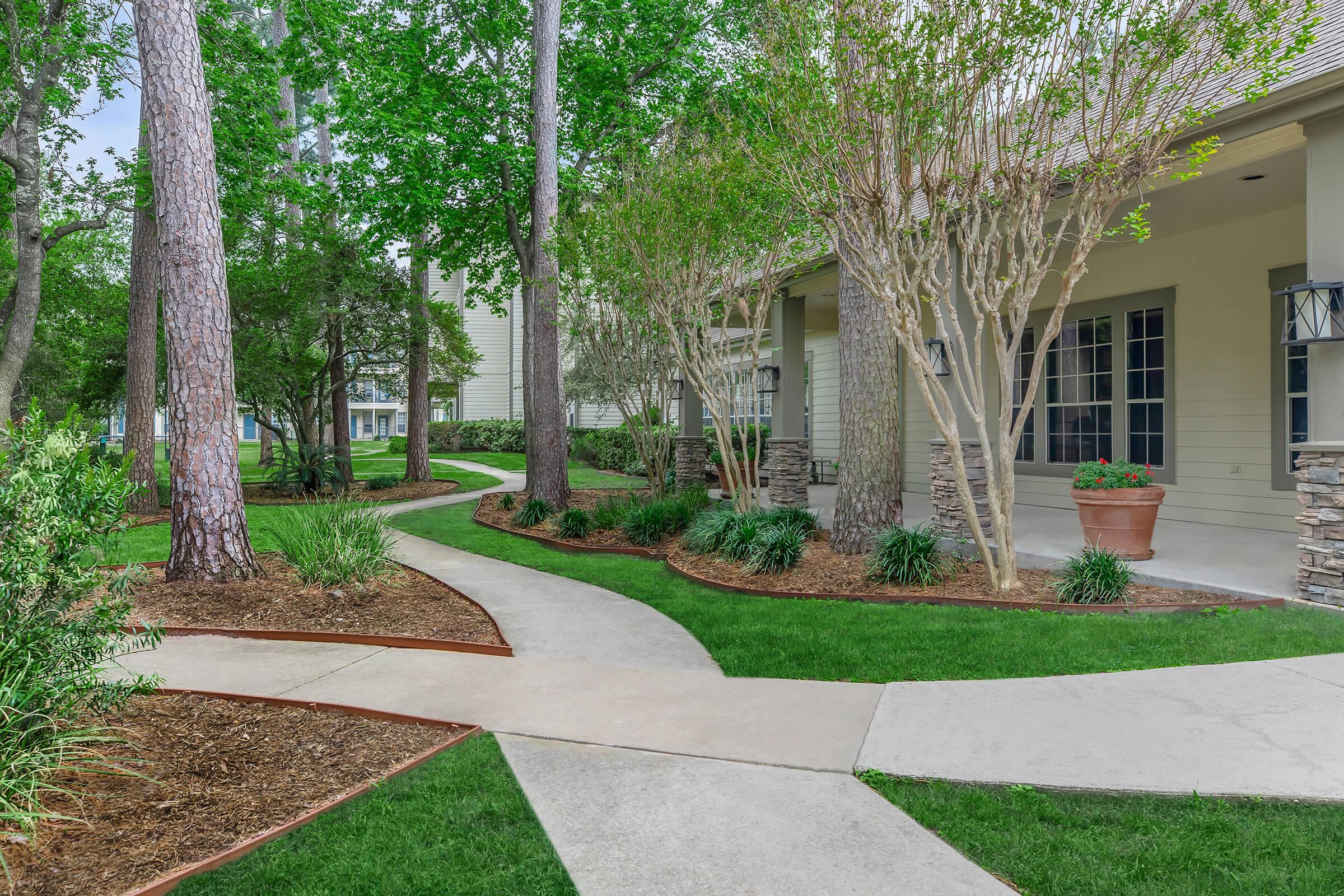 A landscaped pathway winds through a green garden, bordered by trees and shrubs. The path leads to a light-colored building with large windows and a stone accent wall. Fresh grass and potted plants add to the inviting outdoor space.