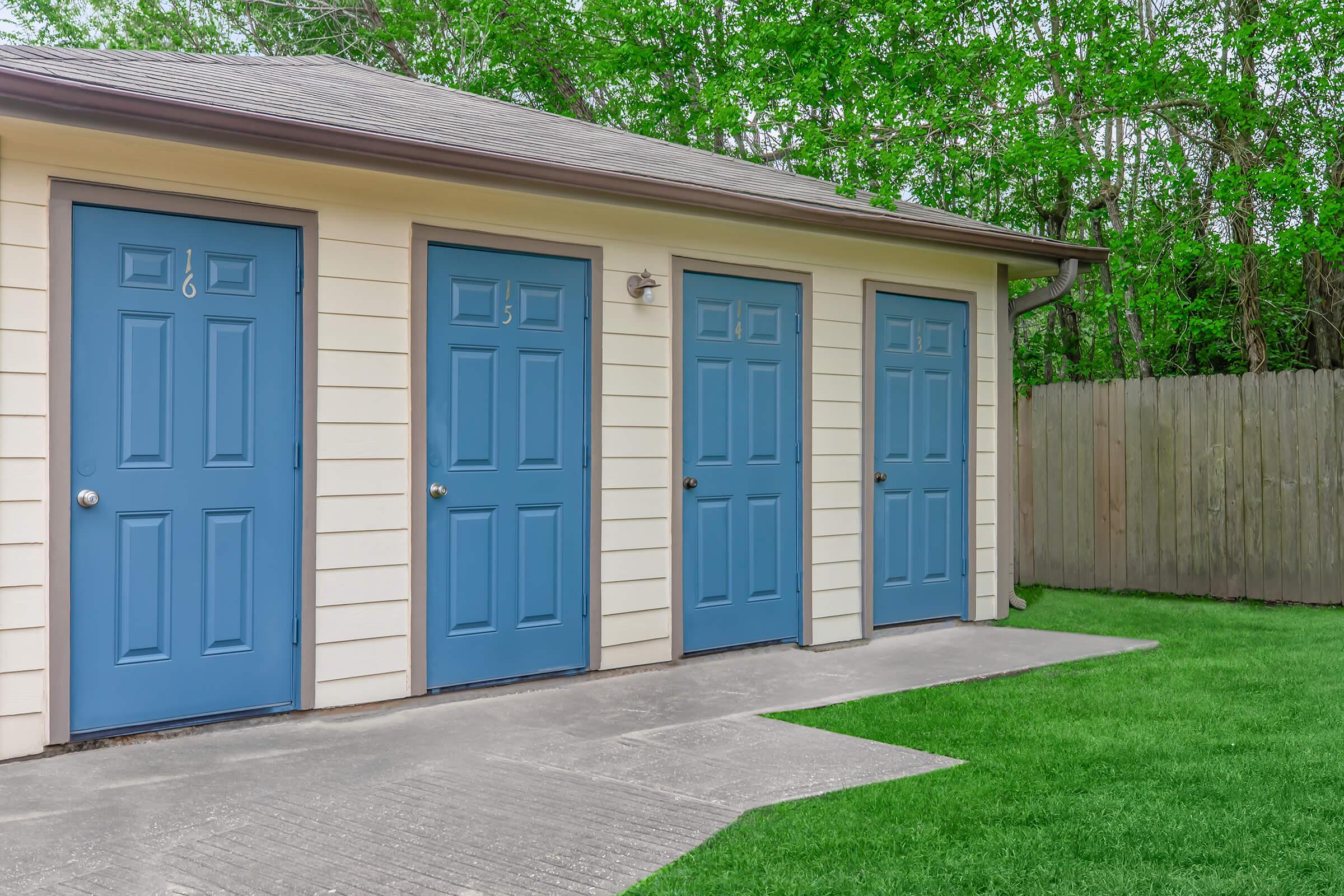 Four blue doors lined up on a light-colored building, set against a backdrop of green trees. The ground is paved with concrete, and there is a well-maintained lawn in front of the building.