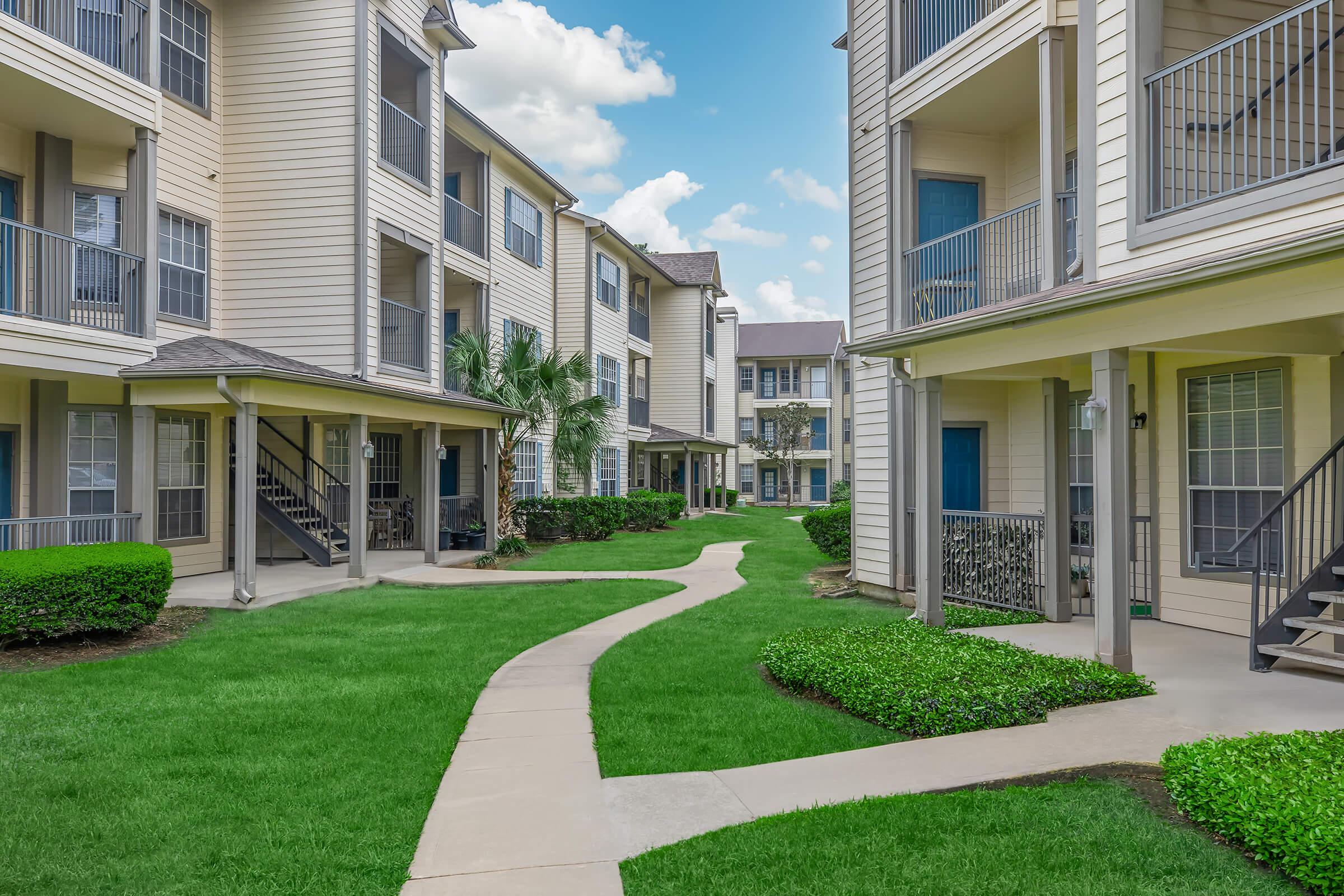 A well-maintained apartment complex featuring beige buildings with balconies. A central pathway winds through lush green lawns and neatly trimmed bushes, leading to the entrances of the apartments. The sky is partly cloudy, adding to the inviting atmosphere of the property.