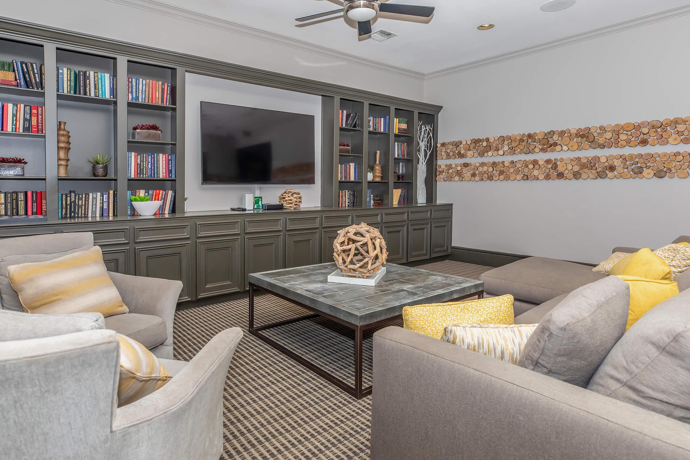 A cozy living room featuring a comfortable seating arrangement with gray sofas and colorful pillows. A central coffee table is surrounded by bookshelves filled with books. There’s a large flat-screen TV on the wall, and decorative wooden wall art adds character to the space. Soft lighting enhances the inviting atmosphere.