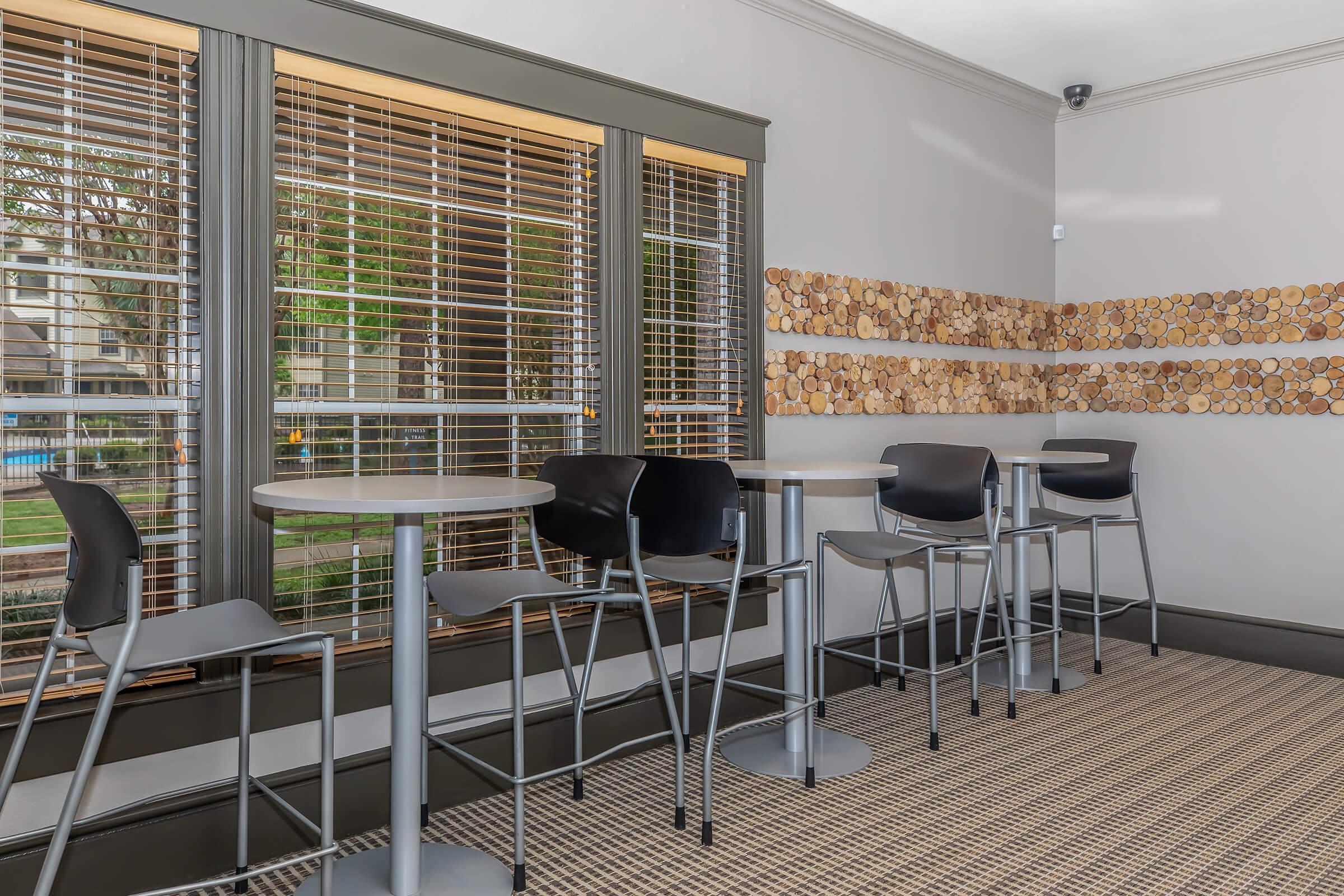 A cozy indoor space featuring high-top tables and black bar stools. Sunlight streams through large windows covered with wooden blinds, highlighting a wooden wall decoration made of circular wood slices. The floor is carpeted in a textured weave, creating a warm atmosphere.