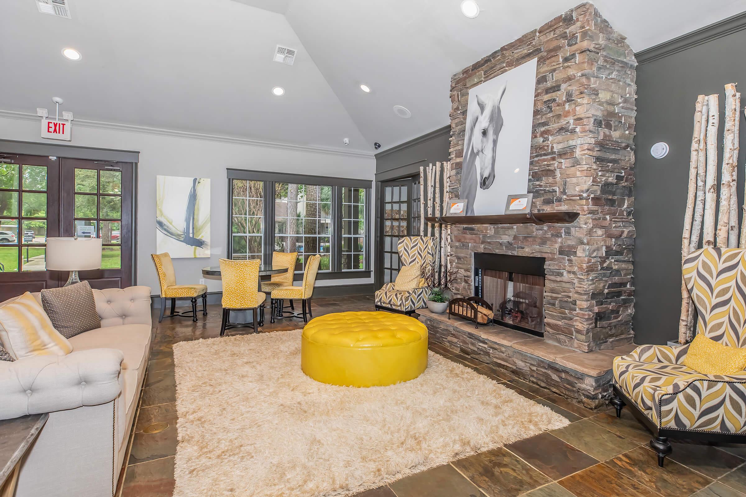 Cozy interior of a modern lounge area featuring a stone fireplace, large yellow ottoman, plush seating, and decorative artwork. The space is illuminated by natural light from large windows and is accented with yellow and gray colors, creating a warm and inviting atmosphere.