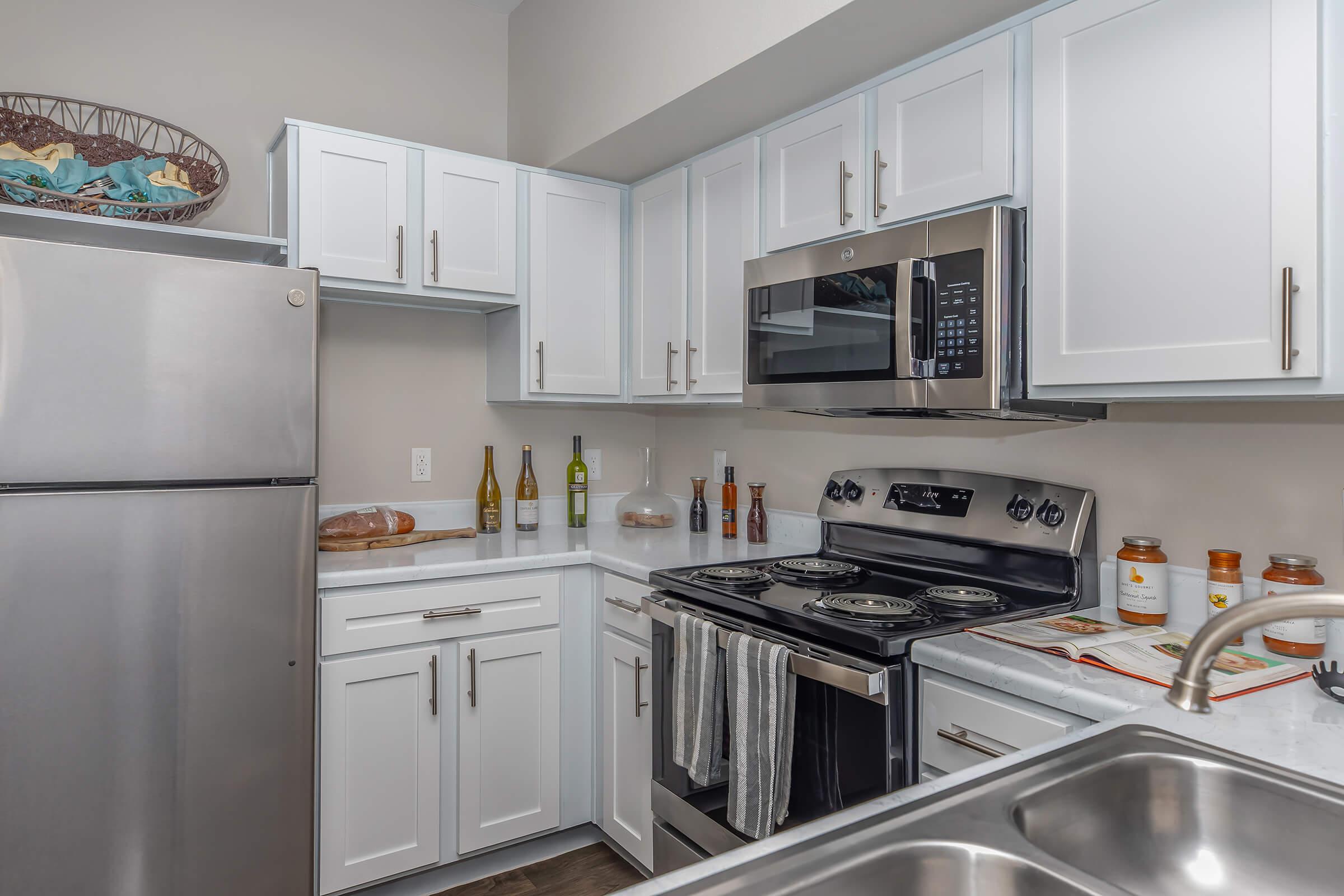 A modern kitchen featuring white cabinetry, a stainless steel refrigerator, and an oven with a microwave above. Countertops are light-colored with various kitchen items, including wine bottles and decorative jars, arranged neatly. The overall design is sleek and inviting, with a warm ambiance.