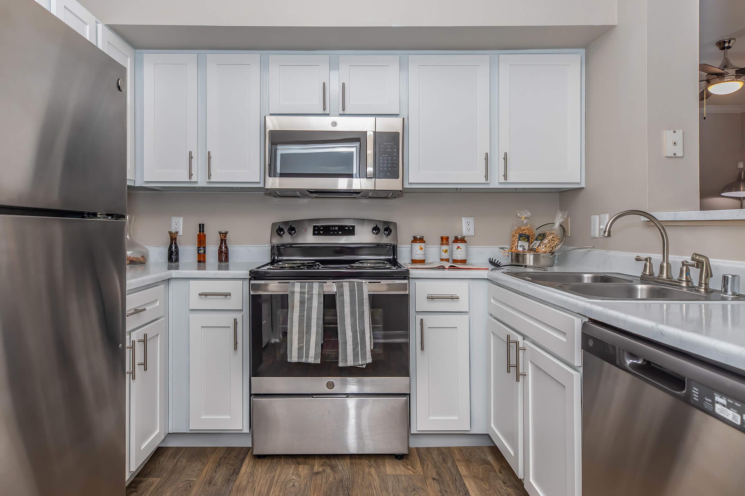 A modern kitchen featuring stainless steel appliances, including a refrigerator, stove, and microwave. The cabinetry is white with silver handles, and there is a double sink with a chrome faucet. Decor items and condiments are placed on the countertop, and the flooring is a wood-like laminate.