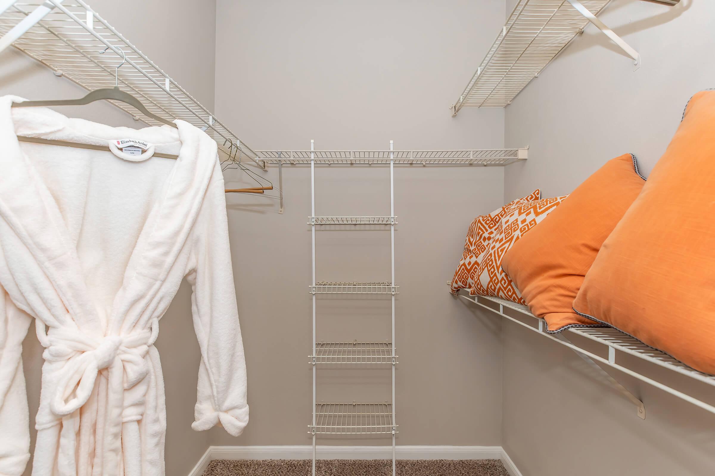 A walk-in closet featuring white wire shelving, a fluffy white bathrobe hanging on a hook, and two orange throw pillows on a shelf. The walls are painted in a light color, and the carpeted floor adds a cozy feel to the space.