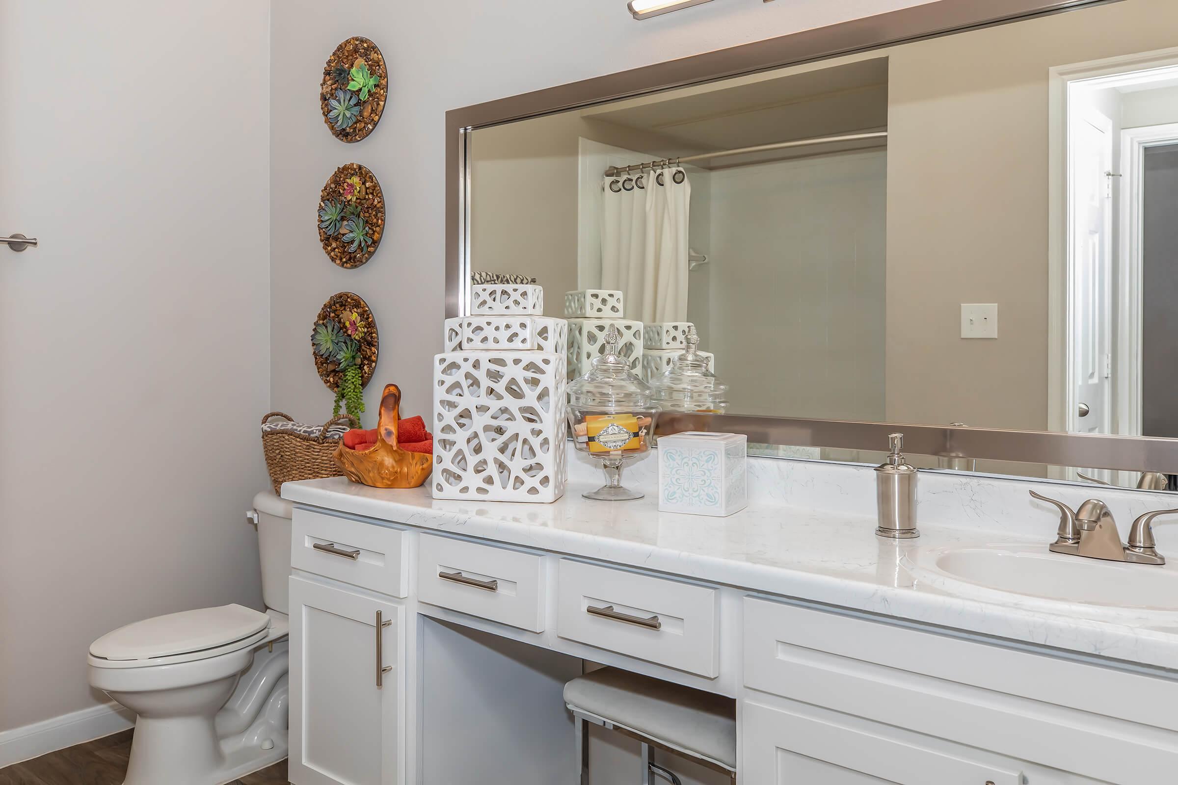 A modern bathroom featuring a white countertop with decorative items, including a glass jar and a small dish. A large mirror reflects the surroundings, and there are circular wall decorations. A clean toilet is visible, along with a shower area behind a frosted glass door.