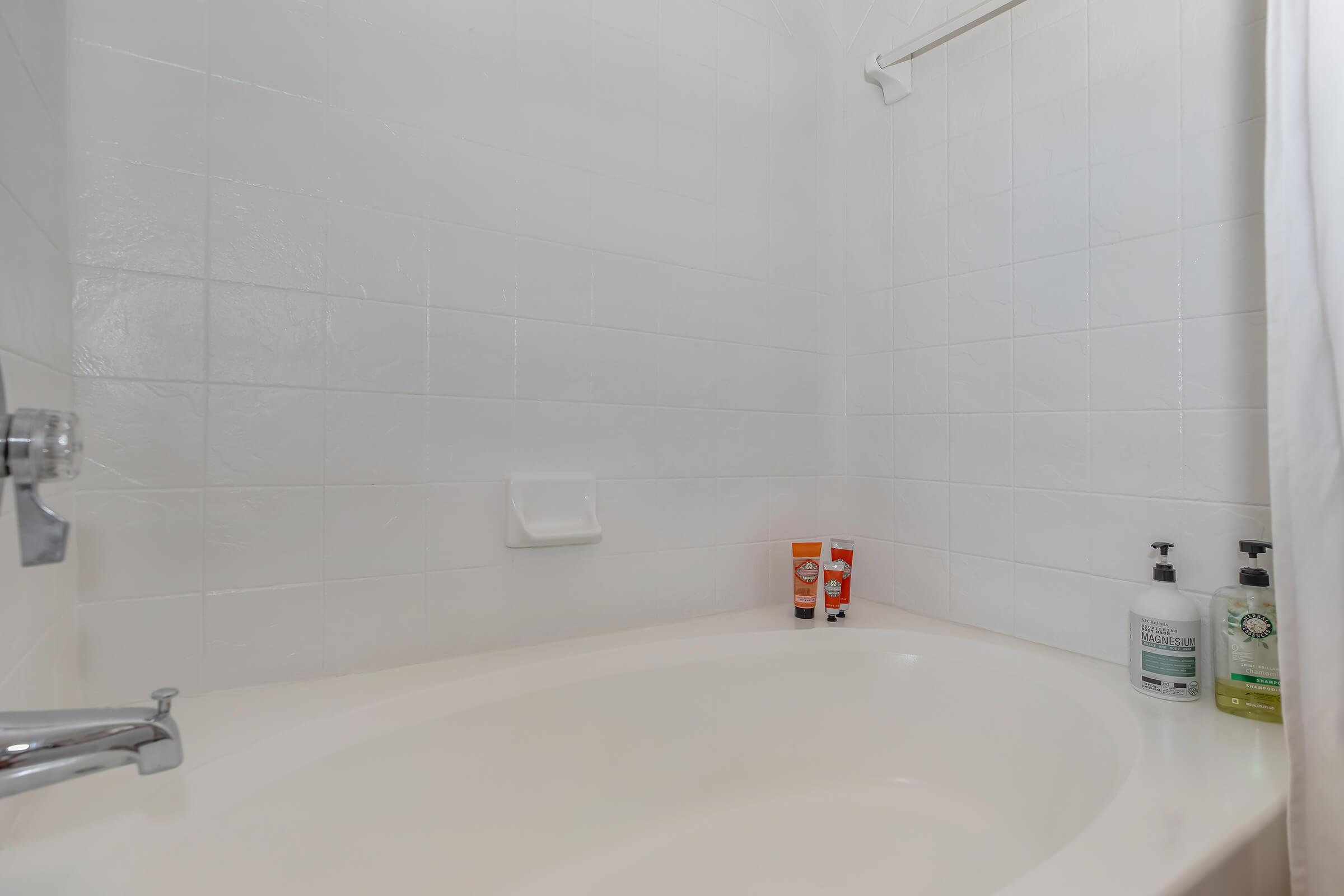 A clean, white bathroom featuring a curved bathtub, white wall tiles, and a shower curtain. On the edge of the tub are three bottles of personal care products, including two tubes and one pump bottle, all neatly arranged. The overall ambiance is bright and fresh.