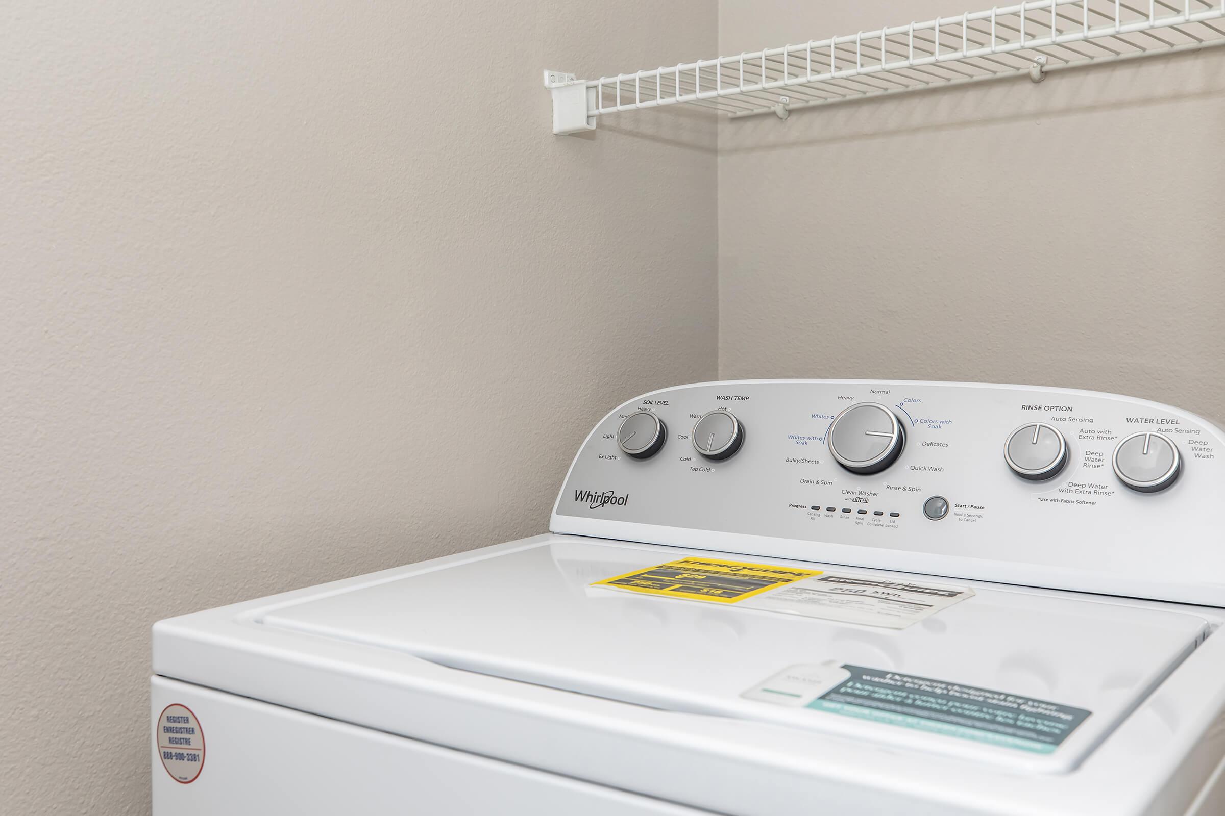 A close-up of a Whirlpool washing machine featuring various control knobs and settings. The machine is placed in a laundry space with a light-colored wall and a wire shelf above. A warning label is visible on the machine's lid, and the overall setting appears clean and organized.