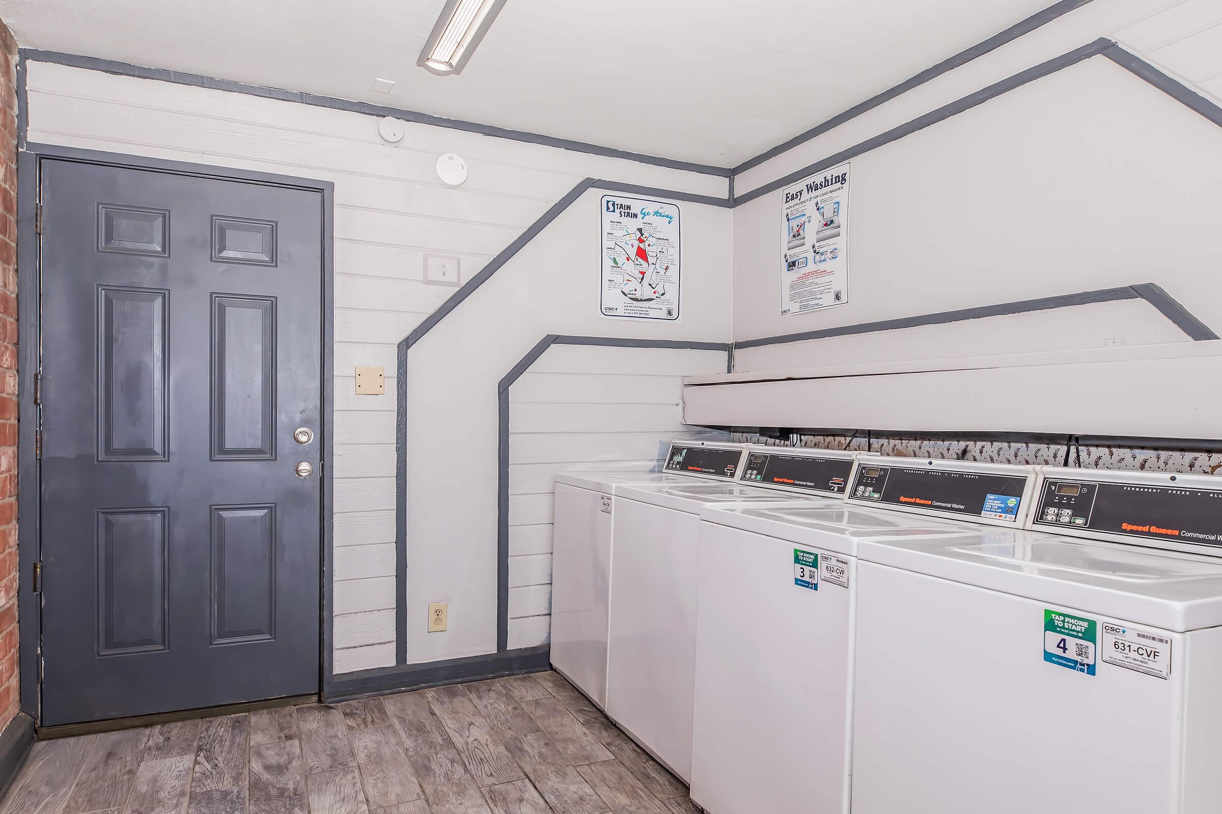 Interior of a laundry room featuring white washing machines and dryers against a gray wall. A dark door is visible, along with a few posters on the wall. The floor is made of light wood-looking tiles, and the space is well-lit with overhead lighting.