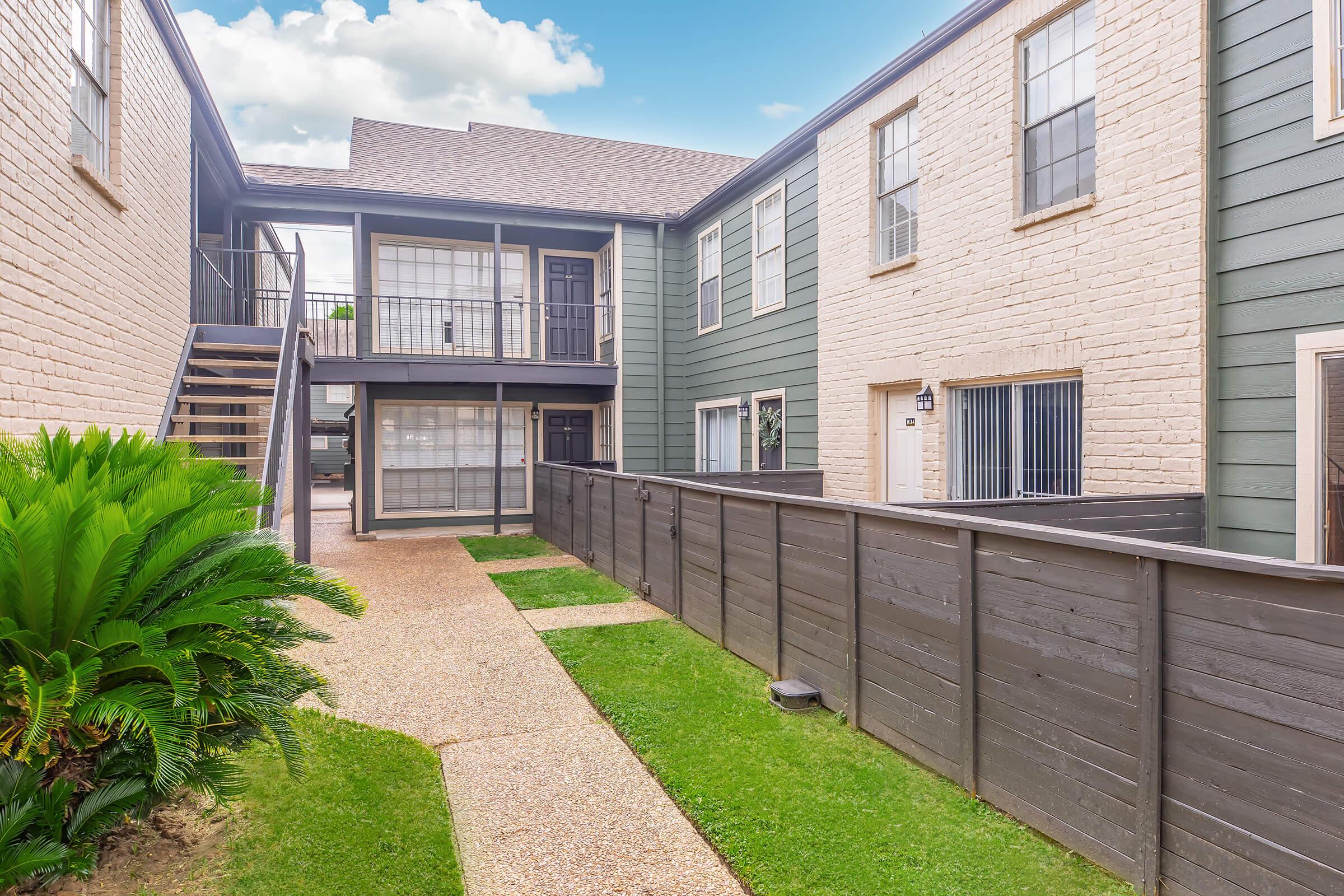 A view of a landscaped apartment complex featuring two residential buildings with a paved pathway between them. The buildings have a mix of light-colored brick and dark siding, and there are lush plants and grass along the walkway. The sky is partly cloudy, giving a bright atmosphere.