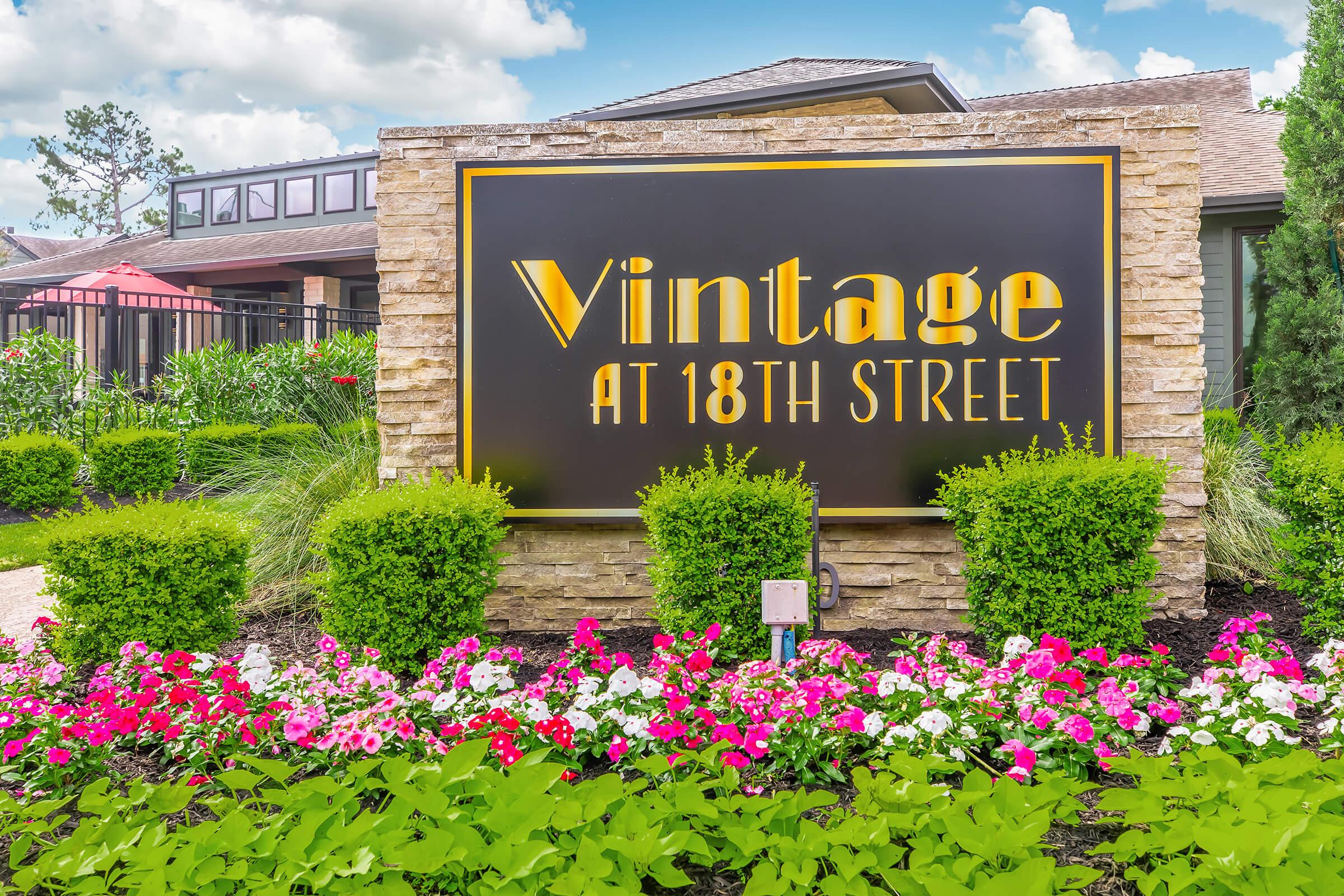 Signage for "Vintage at 18th Street" surrounded by colorful flowers and lush greenery. The sign features a modern font in gold against a dark background, set in a landscaped area with neatly trimmed bushes. Bright blue sky and clouds provide a picturesque backdrop.