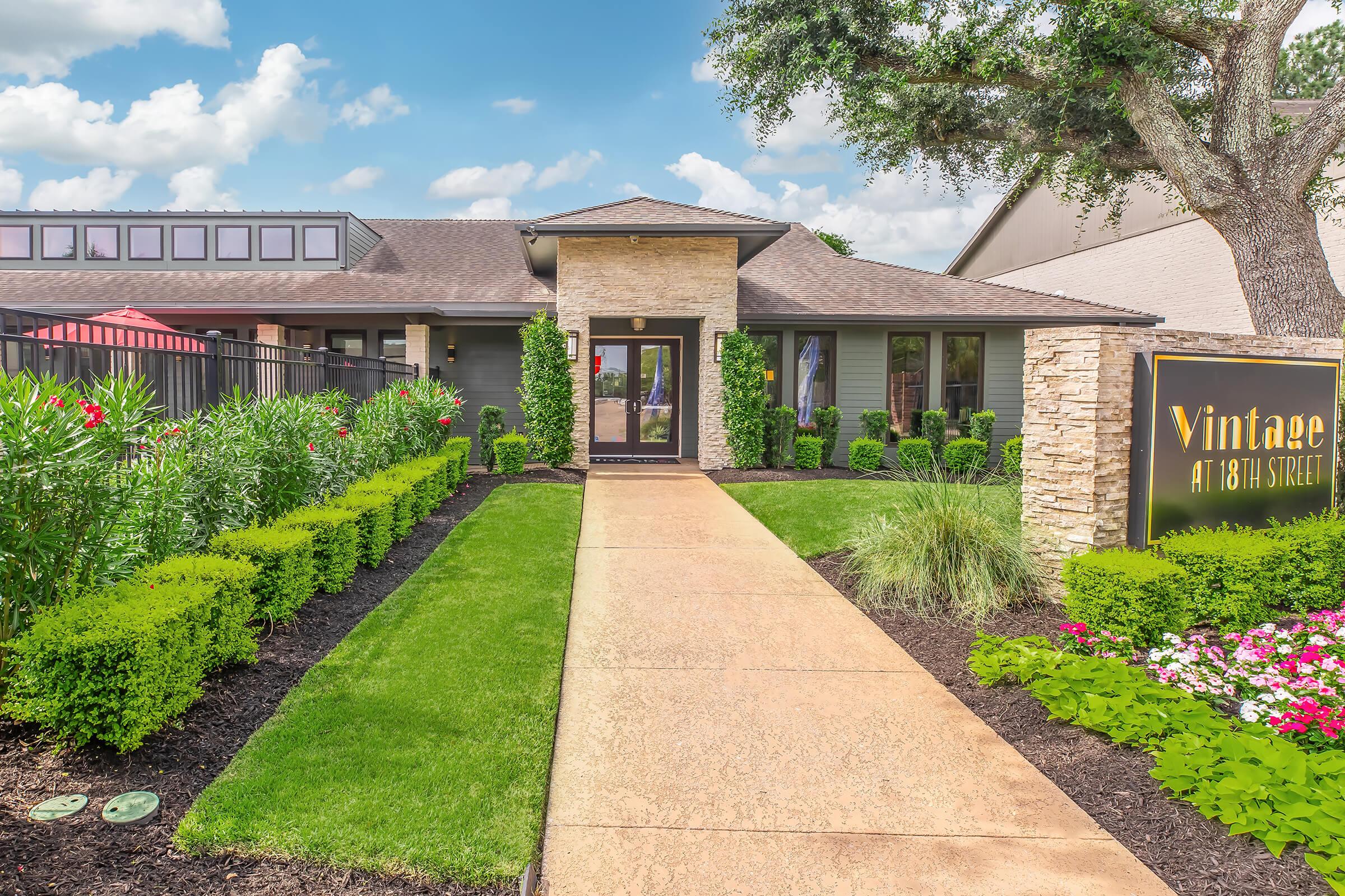 A modern building entrance featuring a stone facade, surrounded by well-manicured lawns and colorful flowers. The pathway leads to a stylish entrance with large glass doors, flanked by lush greenery. A prominent sign displays "Vintage at 18th Street" in a modern font. Bright blue sky with scattered clouds overhead.
