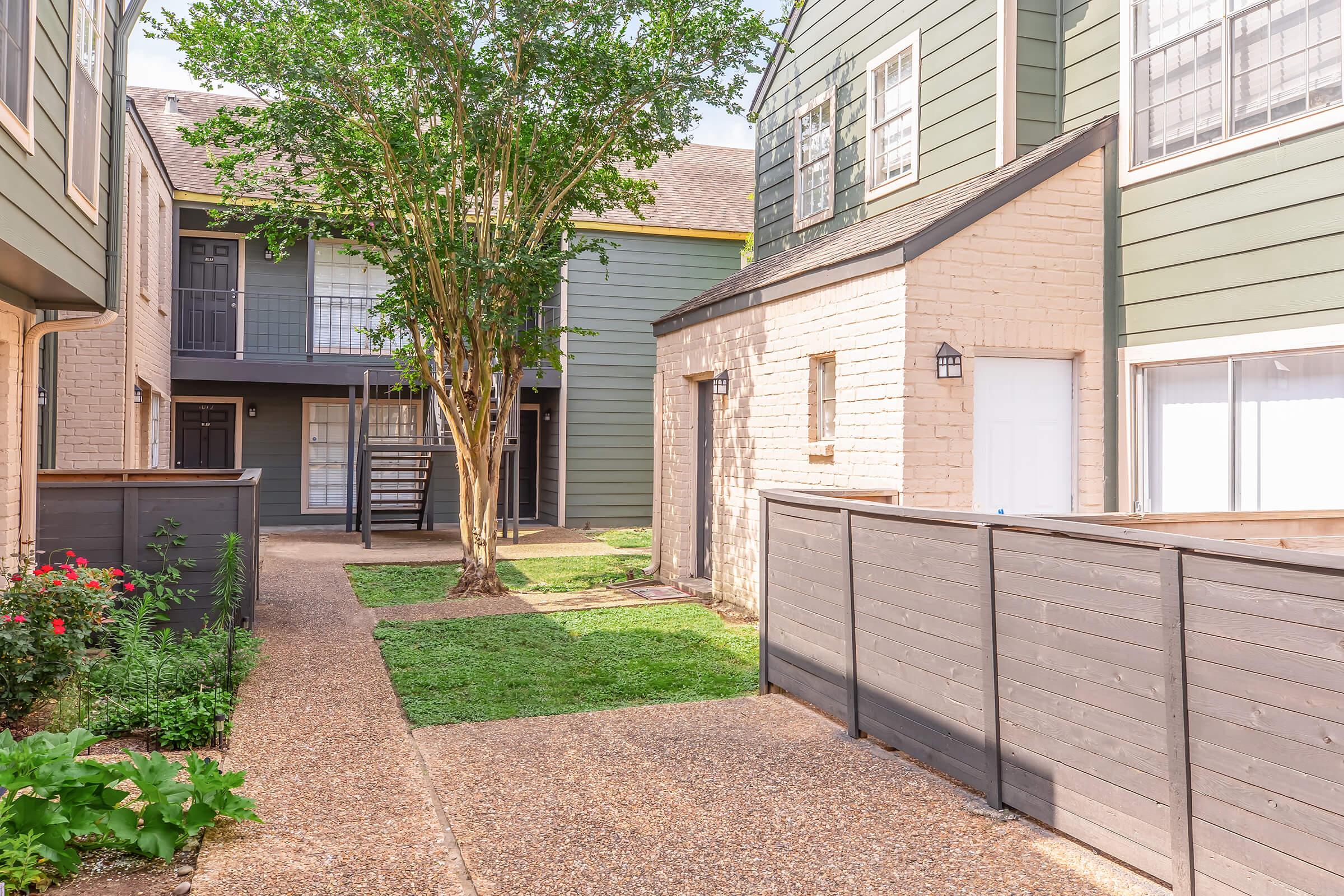 A serene courtyard scene featuring brick and wooden buildings in a residential complex. The pathway is lined with well-maintained grass and flowers, with a small tree providing shade. There are balconies visible from the upper levels, creating a cozy and inviting atmosphere.