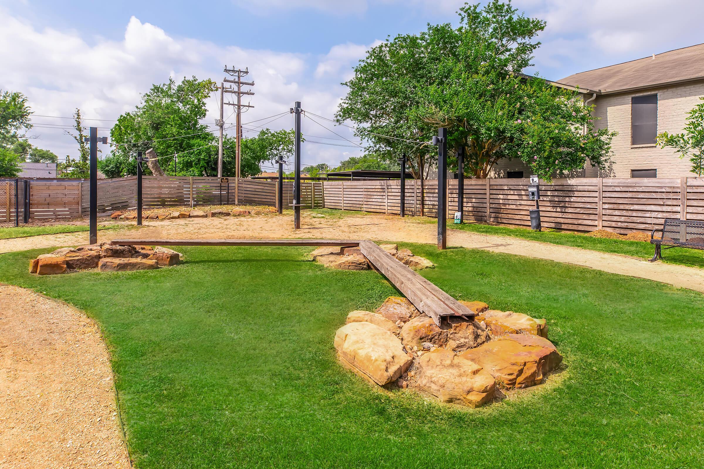 A playground area featuring grassy spaces, rocky formations, and wooden structures. There are several poles, suggesting potential play equipment, and the surrounding area is enclosed with wooden fencing and trees, providing a natural setting. The sky is partly cloudy.