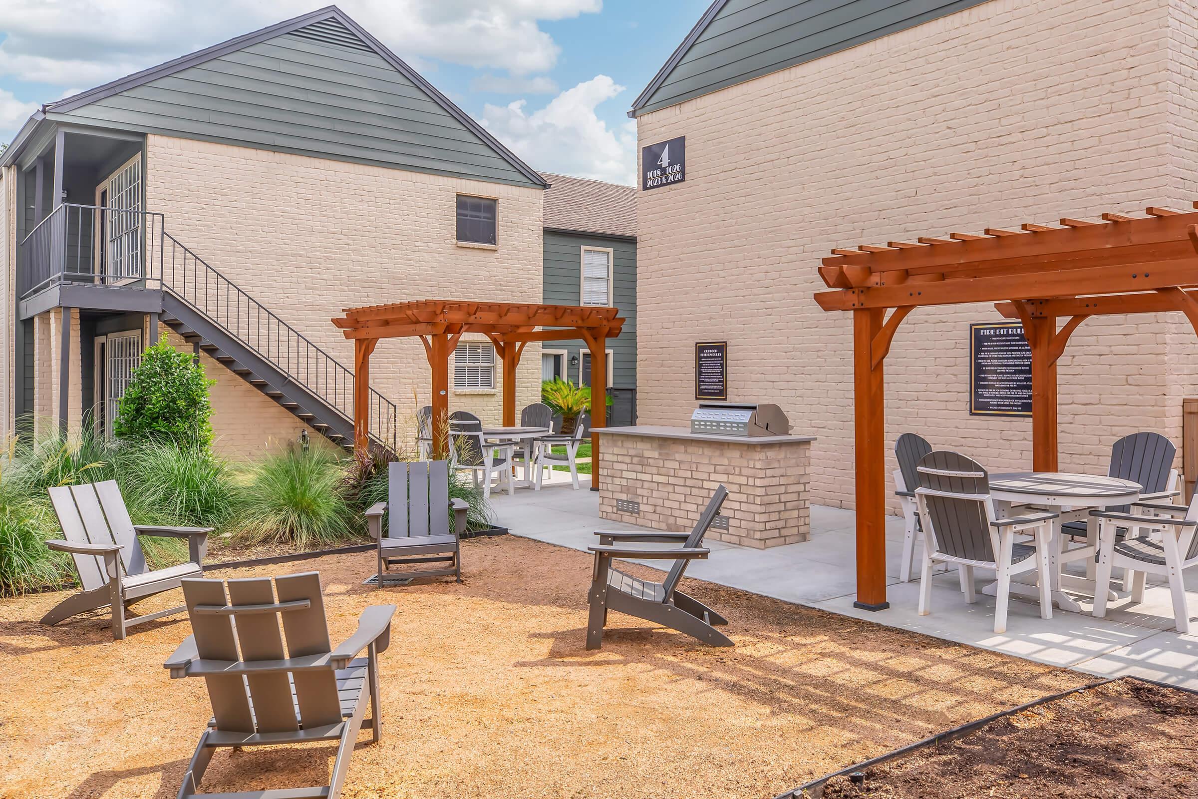 A well-designed outdoor area featuring a gravel landscape with multiple wooden pergolas, outdoor seating with Adirondack chairs, and a communal grill area. Two buildings with light-colored brick exteriors flank the space, surrounded by greenery and under a partly cloudy sky.