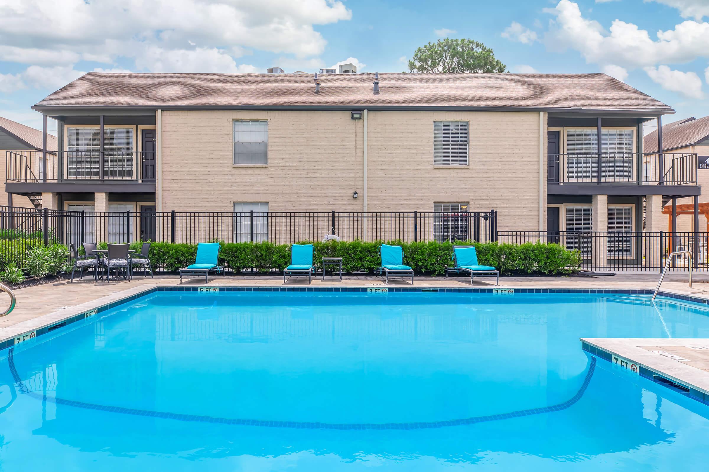 A clear swimming pool with bright blue water is surrounded by lounge chairs. In the background, a two-story building features balconies and large windows. Lush green bushes line the pool area, and the sky above is partly cloudy, creating a relaxing and inviting atmosphere.
