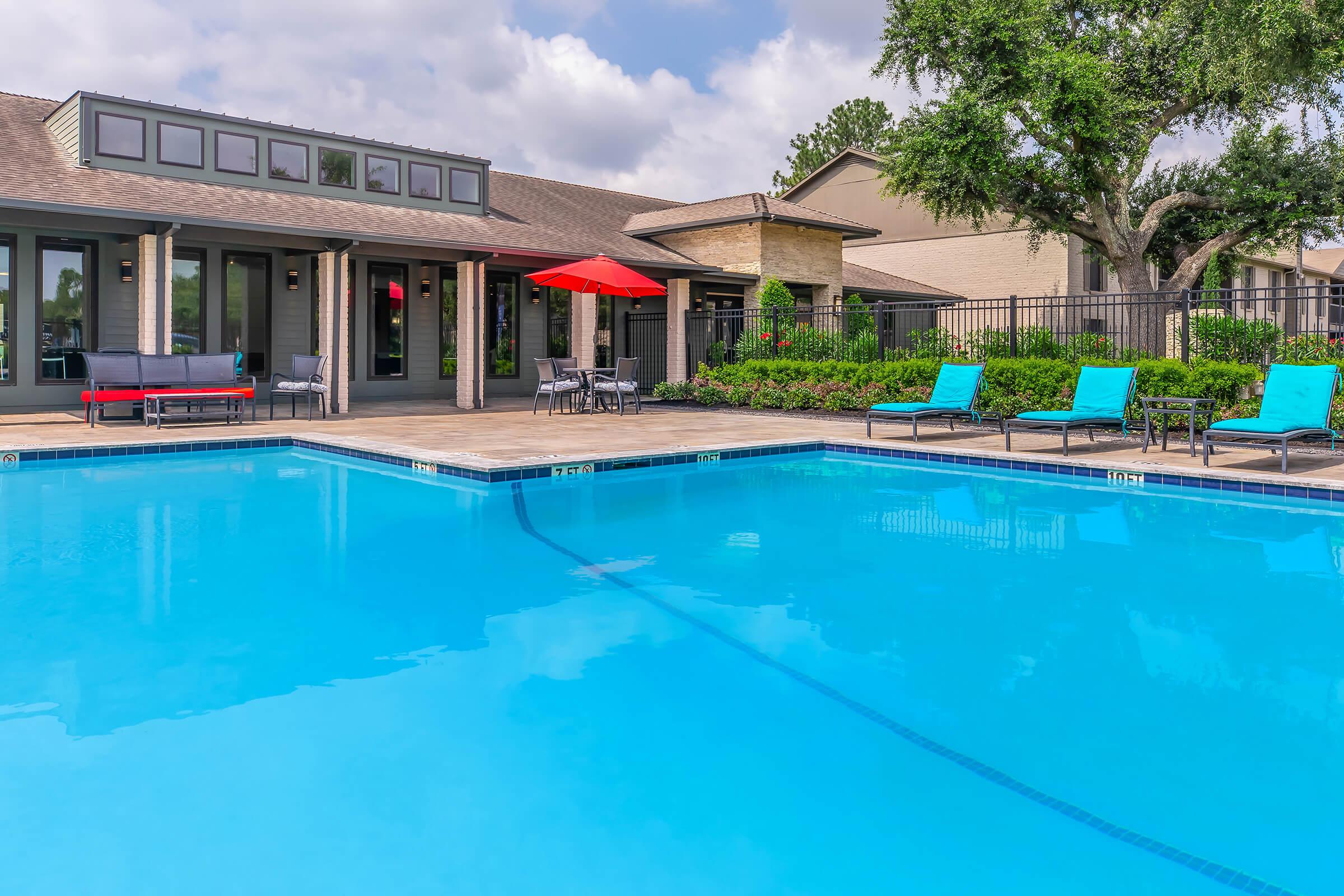 A crystal-clear swimming pool surrounded by lounge chairs and a patio area. There are tables with umbrellas, greenery, and a modern building in the background, creating a relaxing outdoor atmosphere. The sky is partly cloudy, adding to the serene setting.