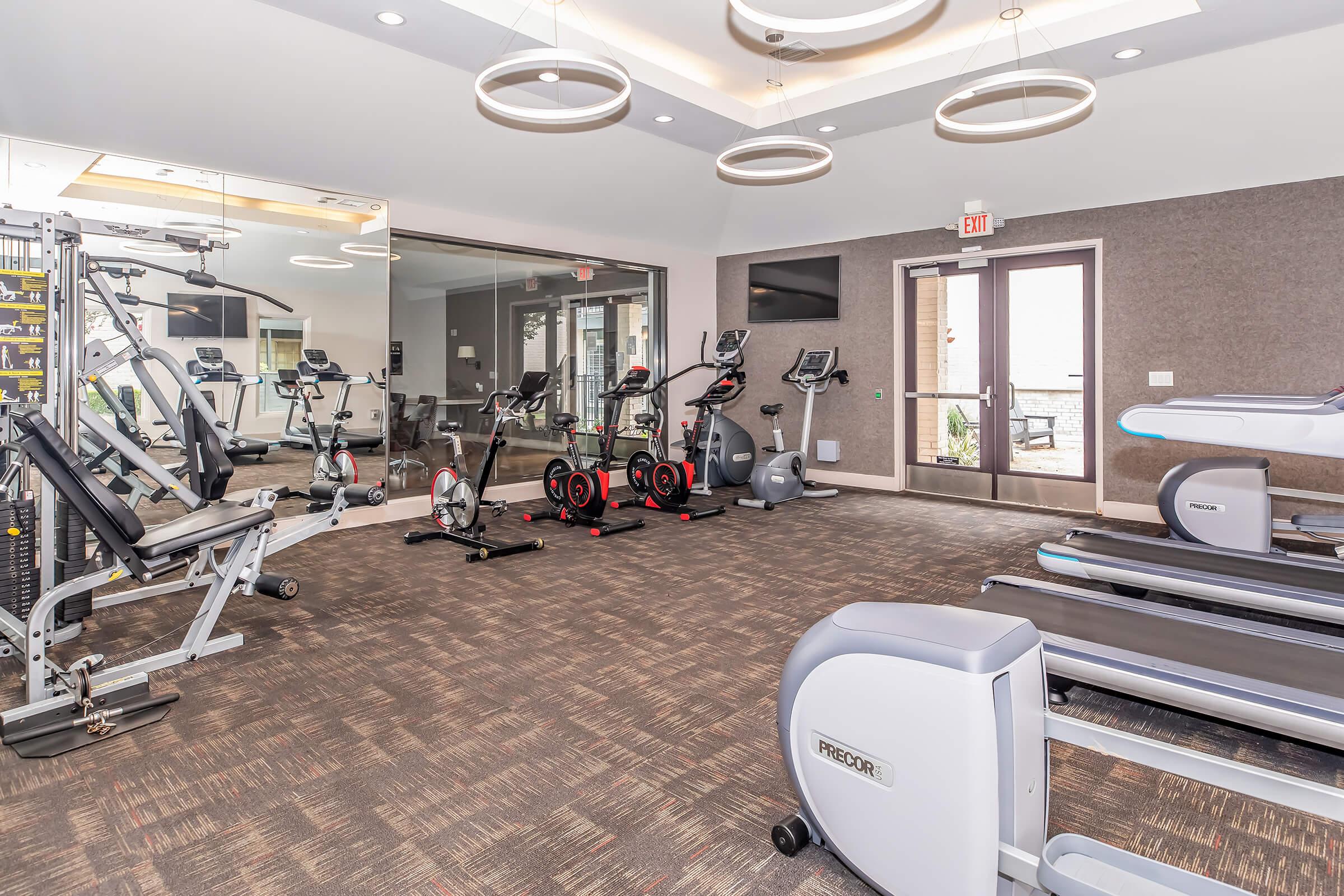 A modern gym interior featuring various exercise equipment, including a weight machine, stationary bikes, and treadmills. The space has large mirrors and contemporary lighting, creating a bright and inviting atmosphere. Large windows provide natural light, enhancing the workout environment.