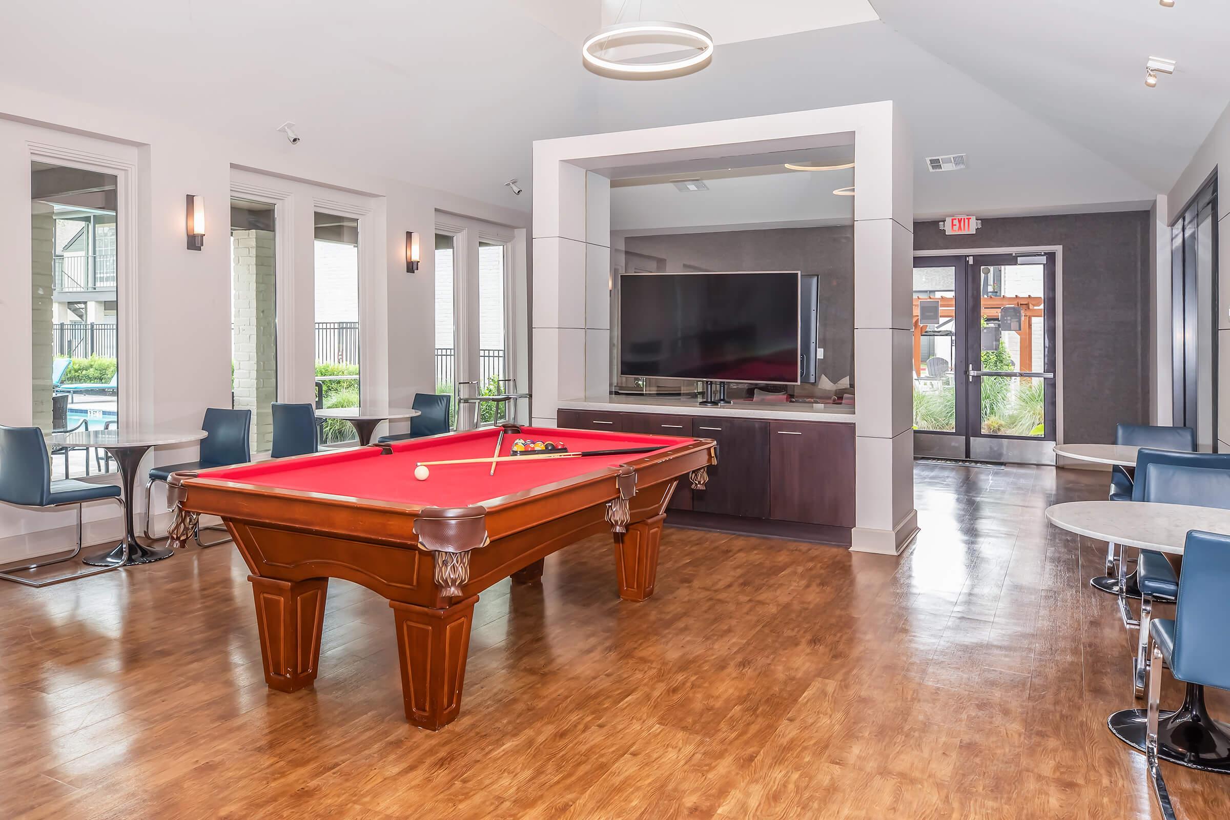 A modern common area featuring a pool table at the center, surrounded by seating options. A large television is mounted on the wall, and floor-to-ceiling windows provide natural light, creating an inviting atmosphere. The room has a contemporary design with warm wood flooring.