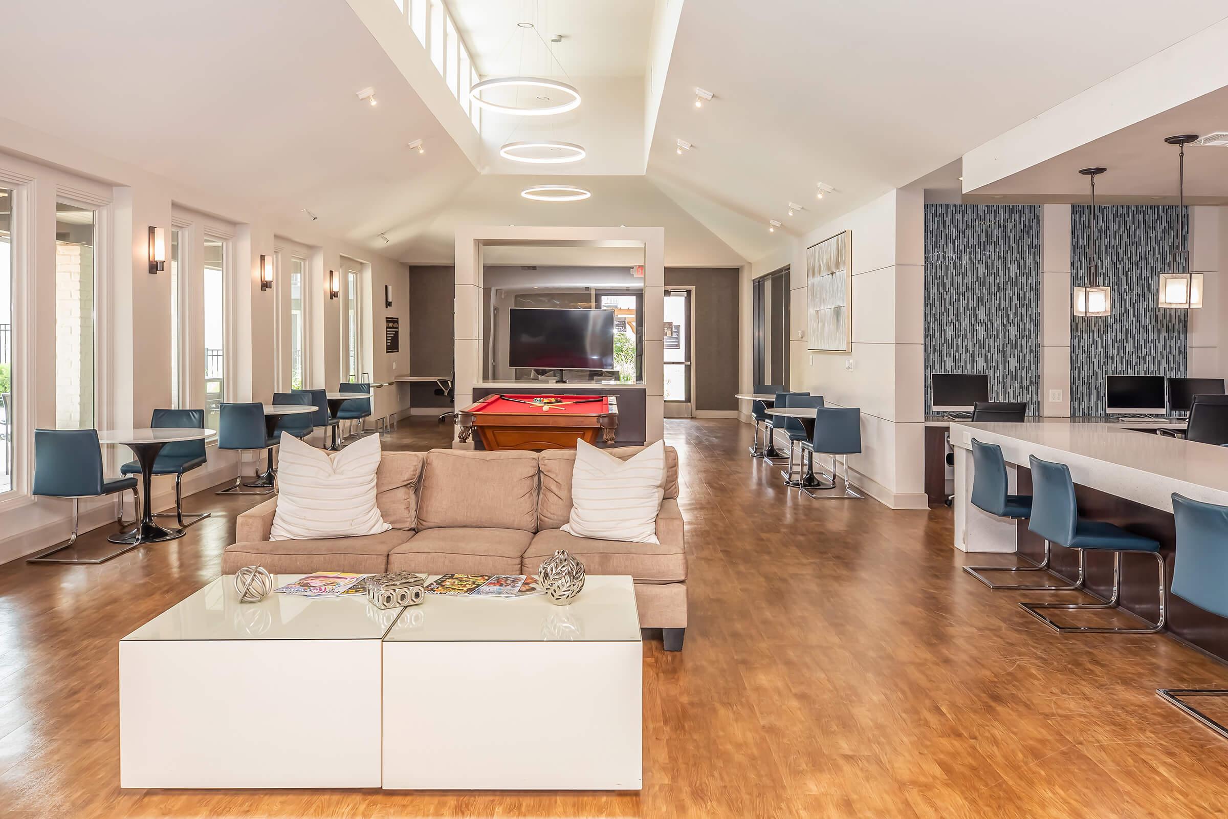 A modern, spacious common area featuring a light-colored sofa with decorative pillows, a glass coffee table, and a pool table. There are several computer stations along one wall and dining tables with chairs, large windows providing natural light, and a contemporary design aesthetic.