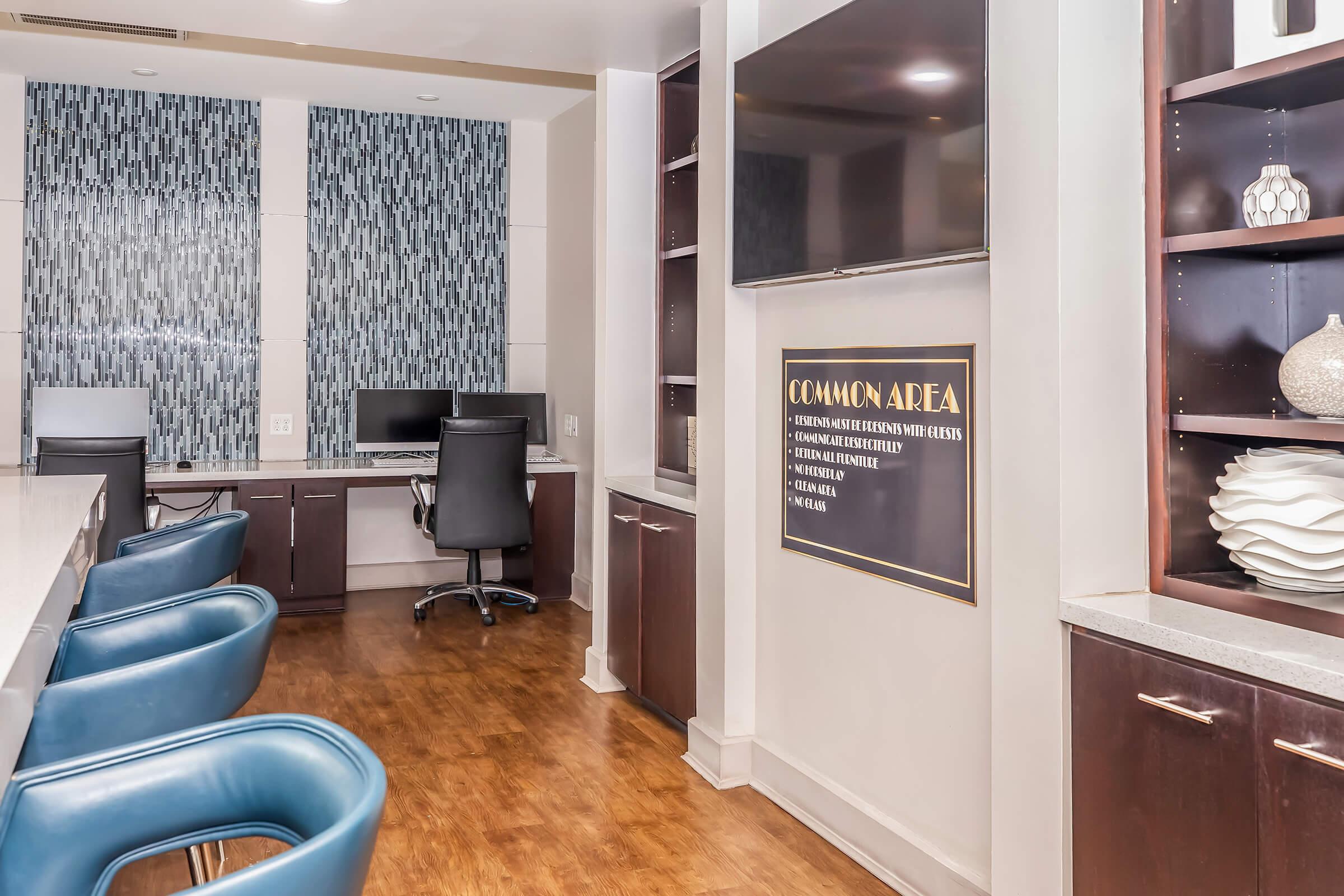 A modern common area featuring a sleek design with a long white countertop and blue bar stools. There are workstations with computers against the wall, and shelves displaying decorative items. A large TV is mounted on the wall, and a sign labeled "COMMON AREA" is prominently displayed.