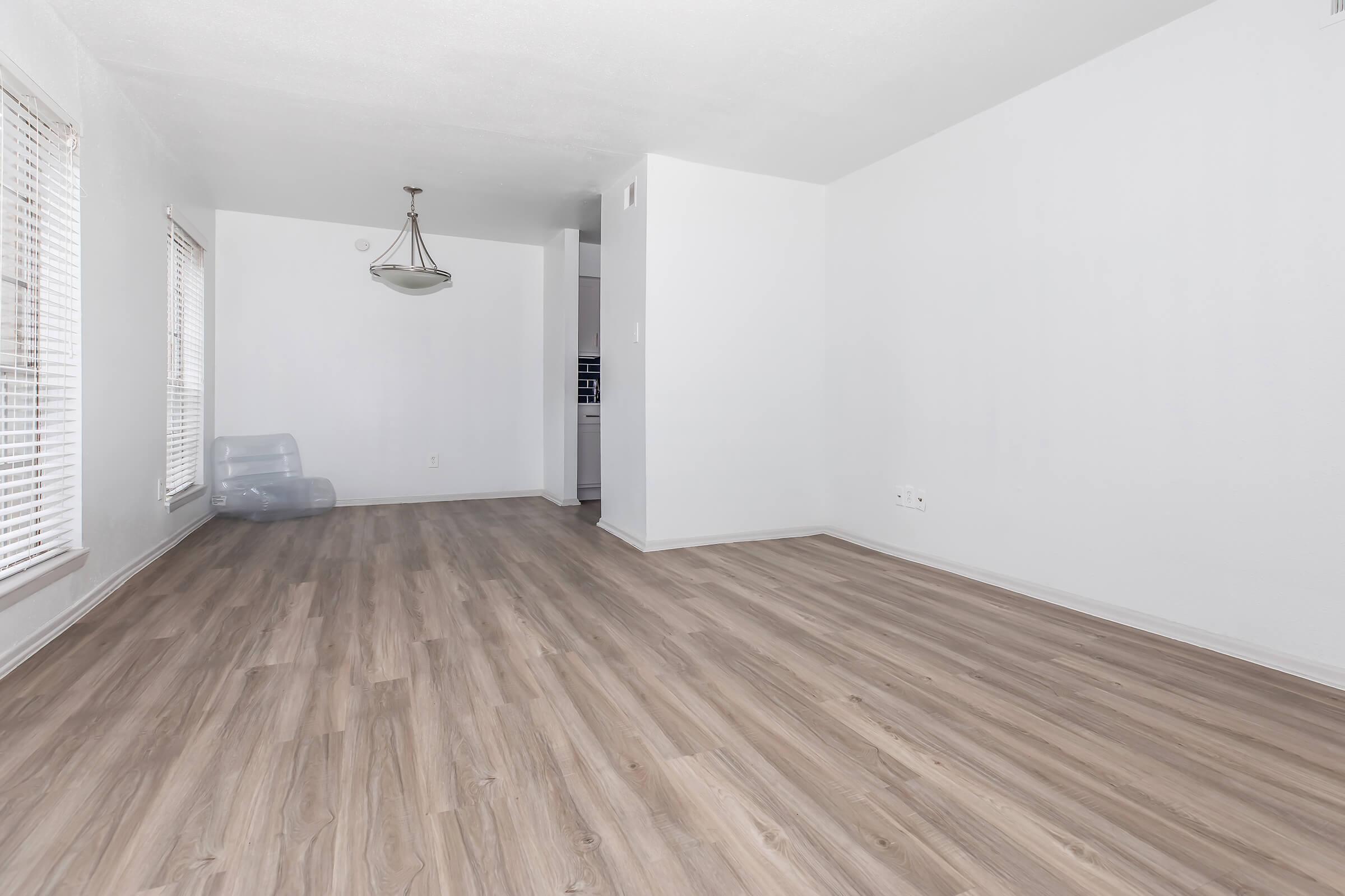 A spacious, vacant room with light-colored walls and laminate flooring. Large windows with blinds let in natural light. A pendant light fixture hangs from the ceiling. A glimpse of a kitchen can be seen in the background. The room is empty, ideal for various furniture arrangements.