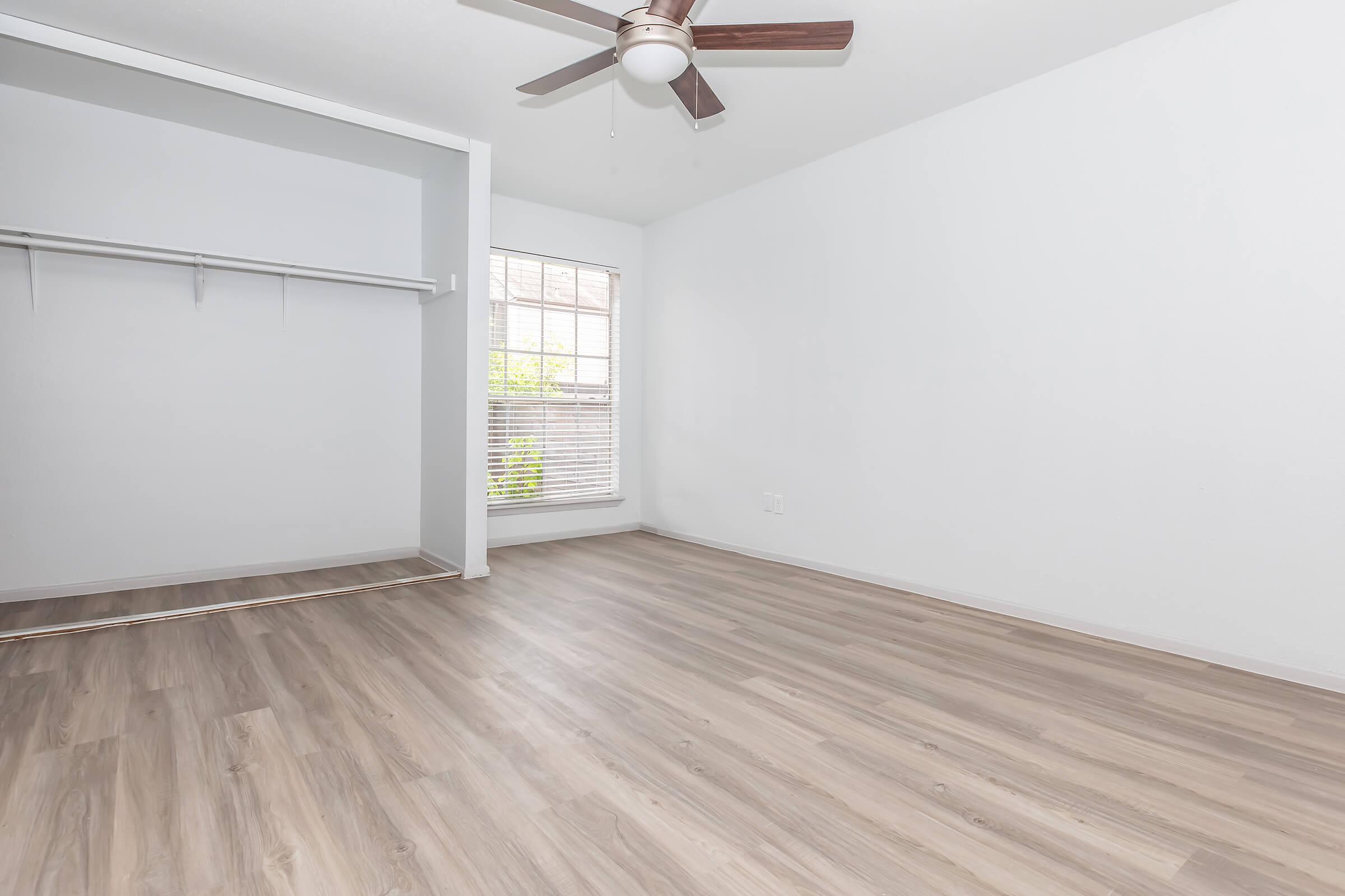 A bright and airy room with light-colored walls and wood-style flooring. The room features a ceiling fan, a window allowing natural light, and an open closet space. The overall ambiance is clean and spacious, suitable for various uses like a bedroom or office.