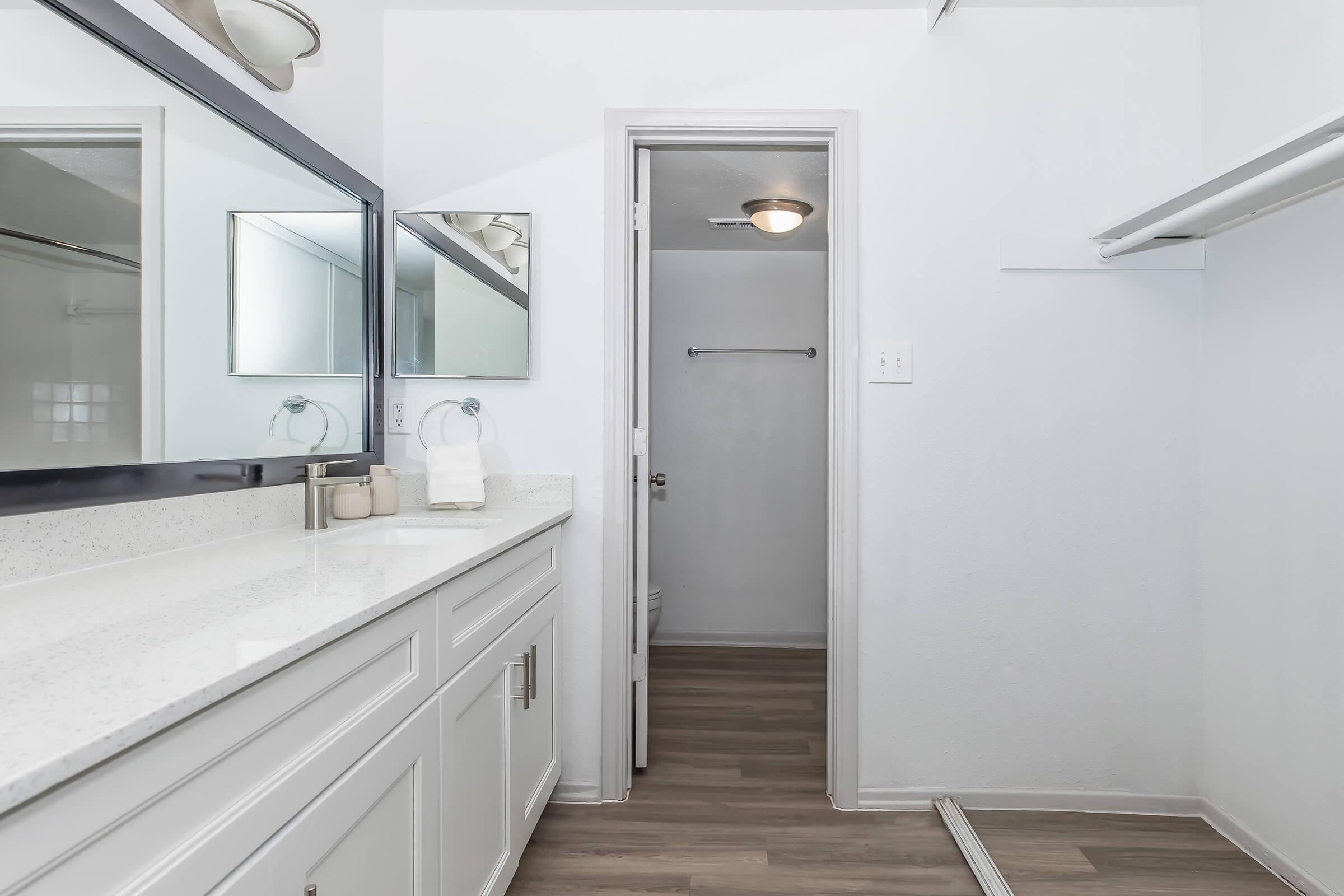A modern bathroom featuring a white vanity with a sink, large mirrors, and neutral-colored walls. There is a doorway leading to a closet or another room, and the floor is covered with light wood-like laminate. The overall design is clean and contemporary.
