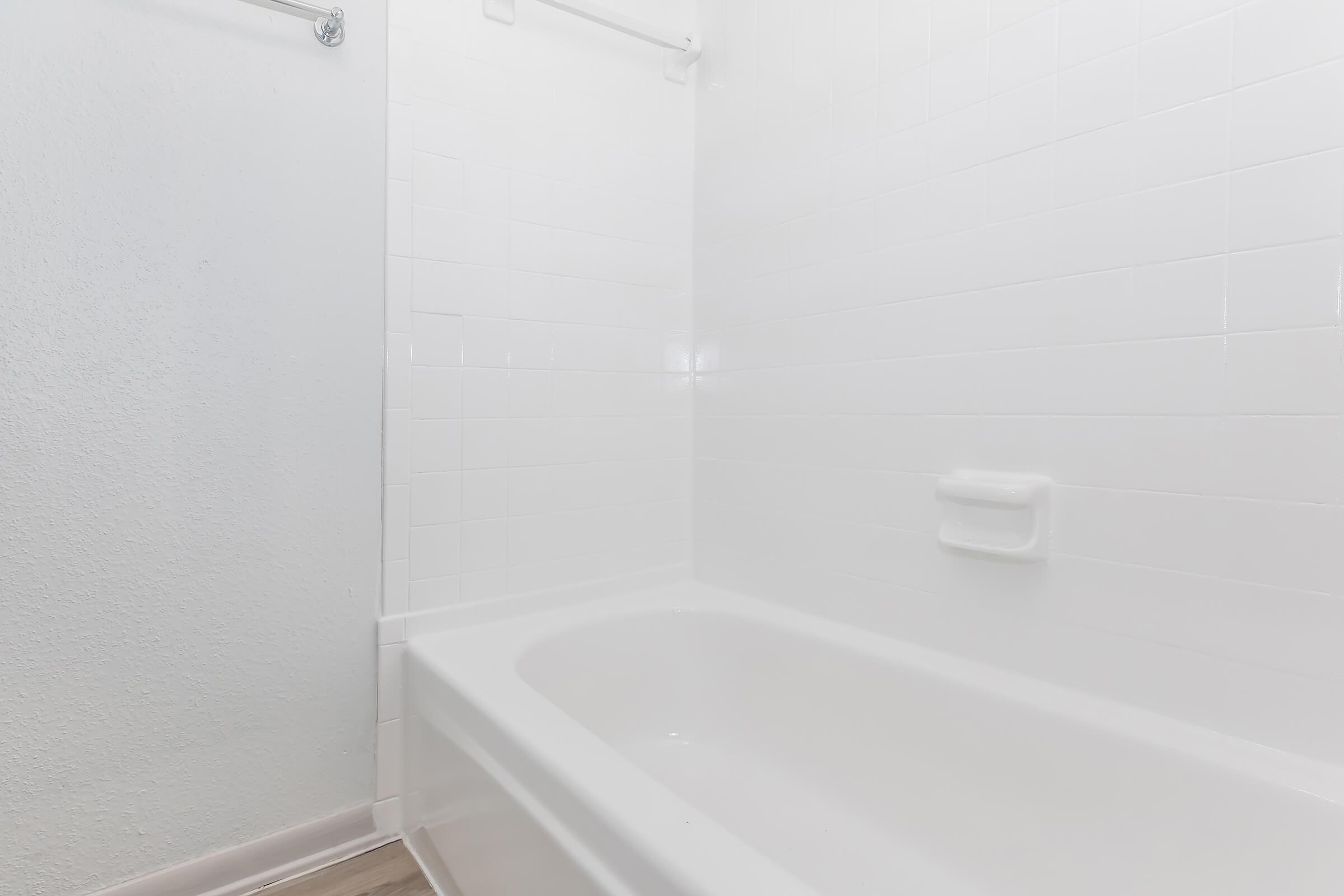 A clean, minimalist bathroom featuring a white tiled wall and a white bathtub. The space is well-lit and has a simple design, emphasizing a fresh and airy atmosphere. There is a towel bar mounted on the wall, adding to the functionality of the space.