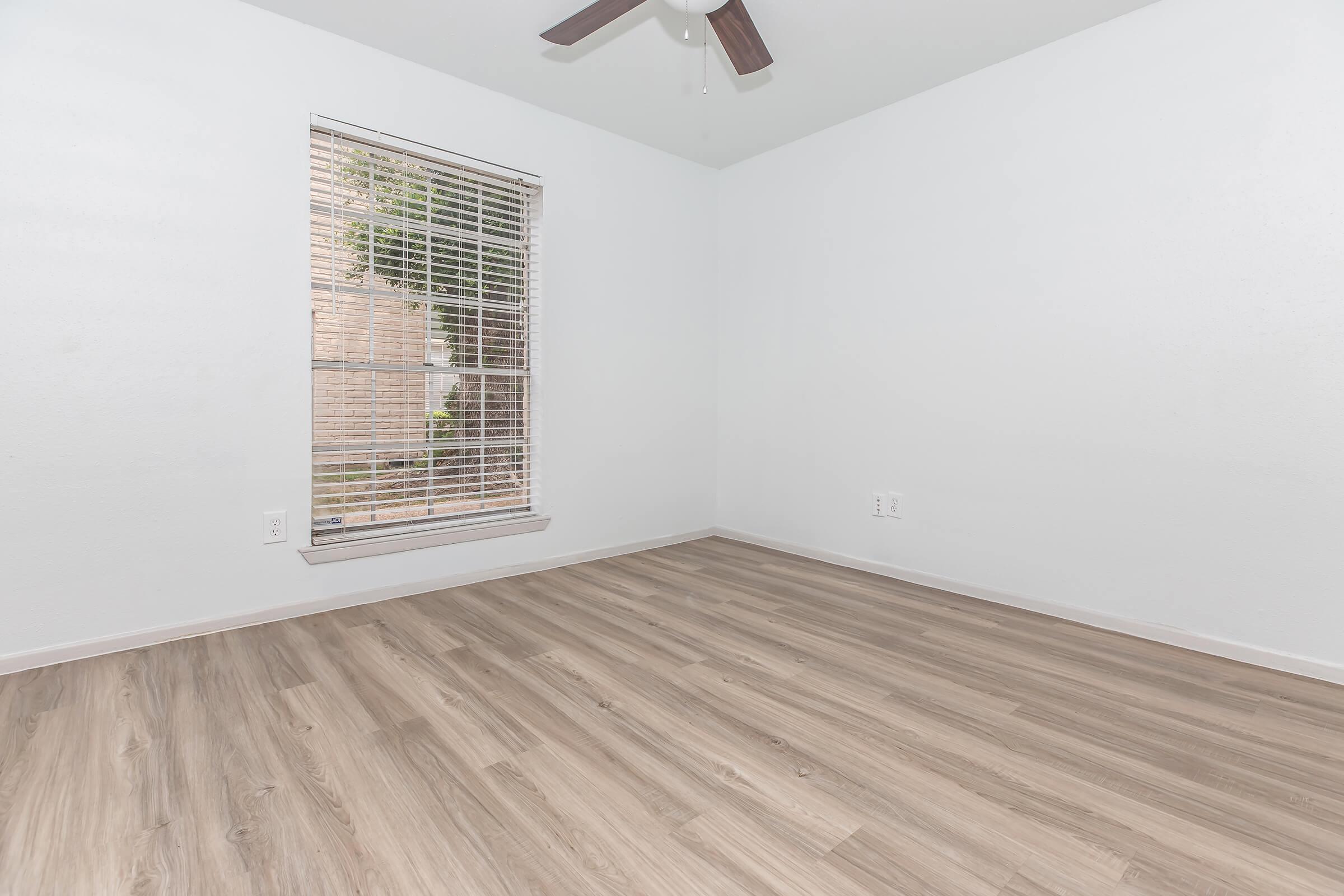 Empty room featuring light-colored walls, a large window with blinds letting in natural light, and a ceiling fan. The floor is covered with light wood-like laminate, creating a clean and spacious appearance.