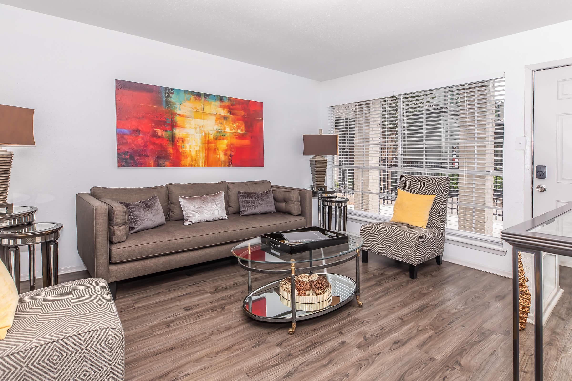 Modern living room featuring a brown sofa with decorative pillows, a glass coffee table with a decorative bowl, and two stylish armchairs. A large colorful abstract painting is mounted on the wall, and there are two table lamps providing warm lighting. The space has hardwood flooring and large windows with blinds.