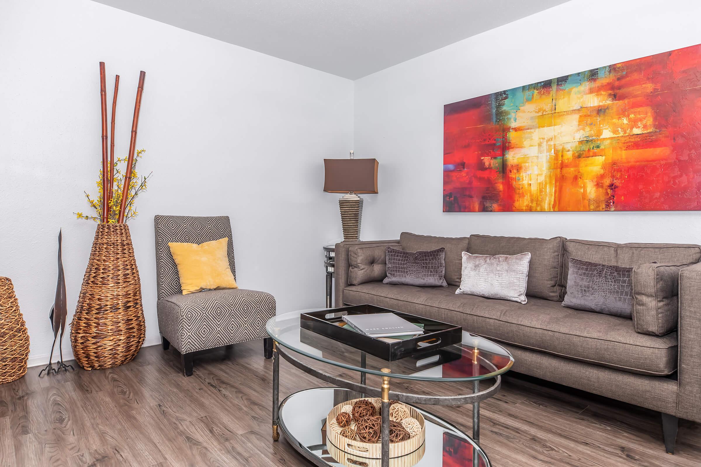 A modern living room featuring a gray sofa with decorative pillows, a glass coffee table displaying decorative objects, a patterned chair, and a tall woven basket. A vibrant abstract painting hangs on the wall, complemented by a lamp and decorative branches in another basket. The floor is covered in wood-like laminate.