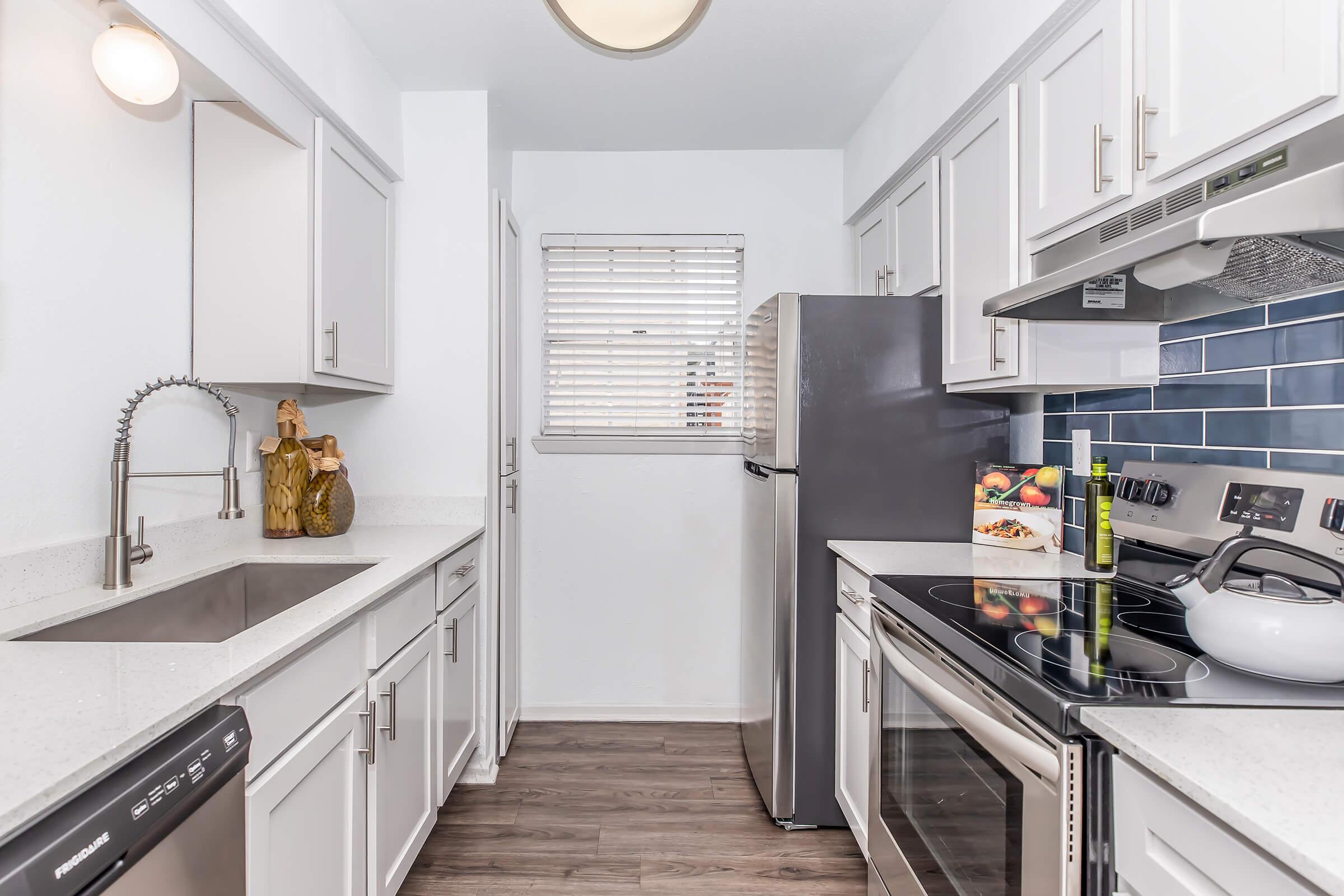 Modern kitchen featuring white cabinets, a stainless steel refrigerator, an electric stove, and a sleek sink with a stylish faucet. The walls are adorned with a blue tile backsplash, and there is a window with blinds allowing natural light. A fruit basket is placed on the counter, adding a homey touch.