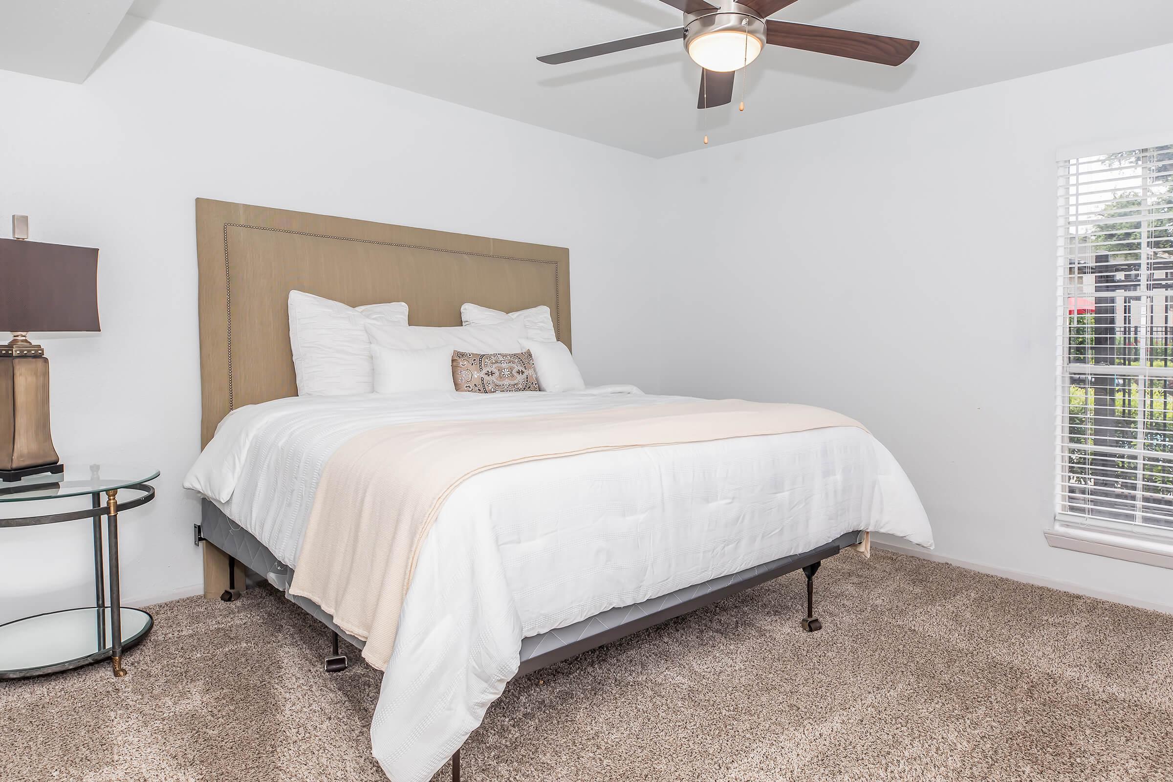 A cozy bedroom featuring a queen-sized bed with white bedding and decorative pillows, a bedside table with a lamp, and a window with blinds letting in natural light. The room has light-colored walls and plush carpeting, creating a warm and inviting atmosphere.