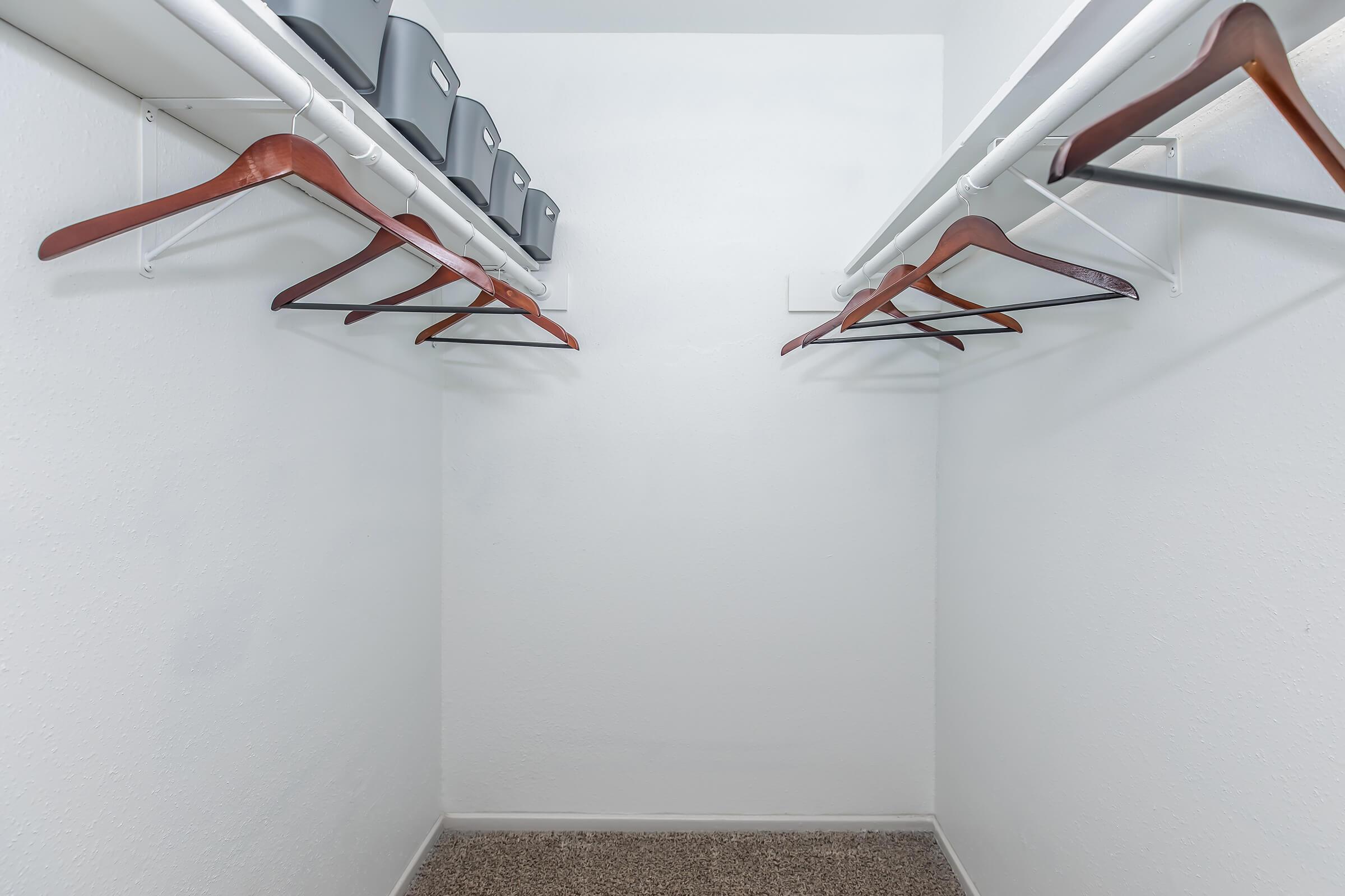 A clean, organized closet featuring empty wooden hangers on two white rods. Above the hangers, there are several gray storage bins neatly arranged. The walls are white, and the floor is carpeted in a neutral color, creating a spacious and tidy appearance.