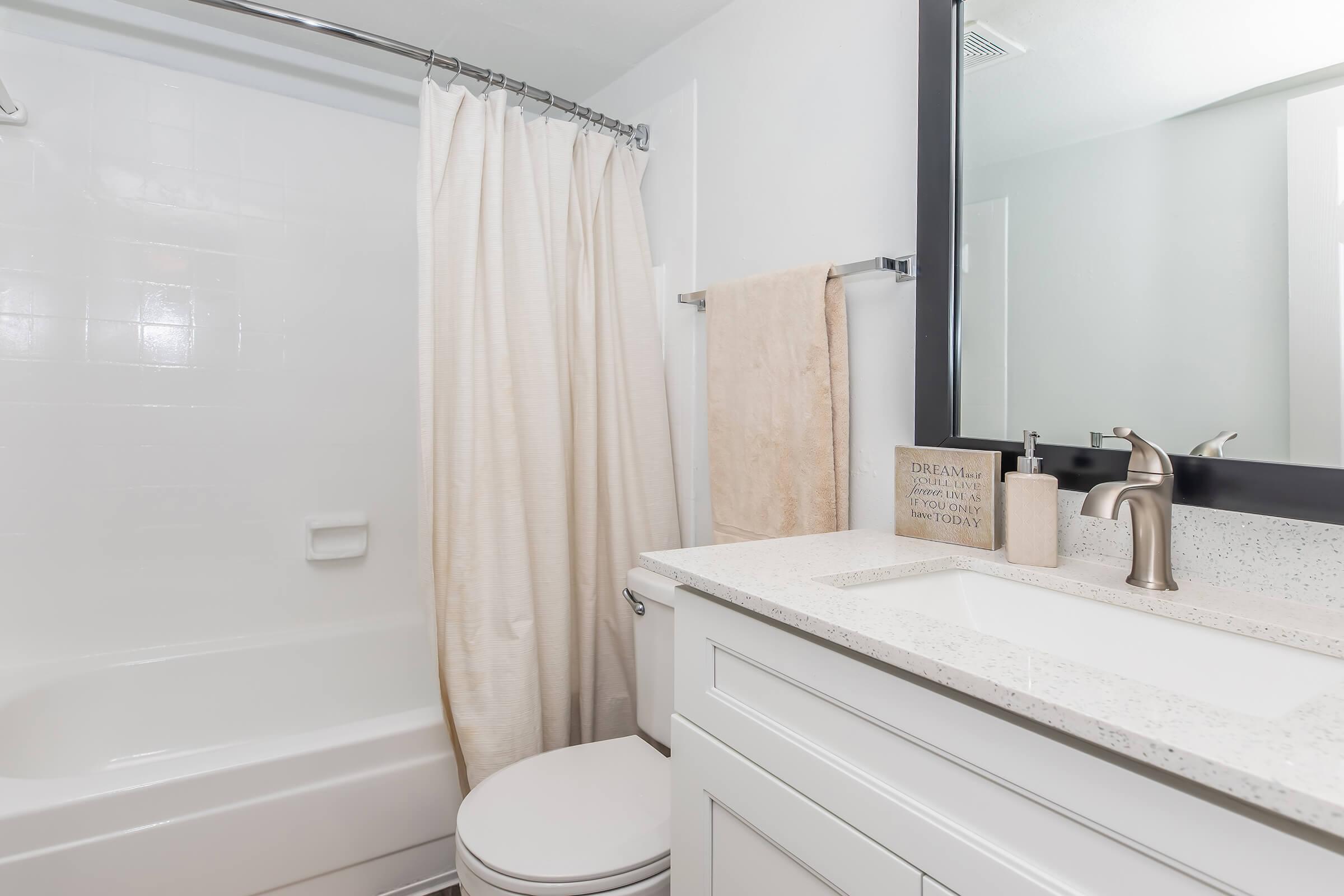A clean and modern bathroom featuring a bathtub with a shower curtain, a sink with a countertop, a mirror above the sink, and neatly arranged towels. The decor is simple and includes a decorative sign on the counter. The overall color scheme is light and airy, creating a fresh atmosphere.