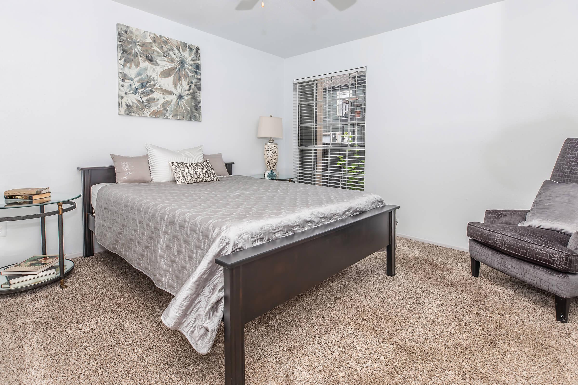 A cozy bedroom featuring a queen-sized bed with a gray comforter, two decorative pillows, and a nightstand with a lamp. A chair sits in the corner, and a round side table holds a few books. Natural light enters through a window with blinds, and the carpet is a light brown shade.