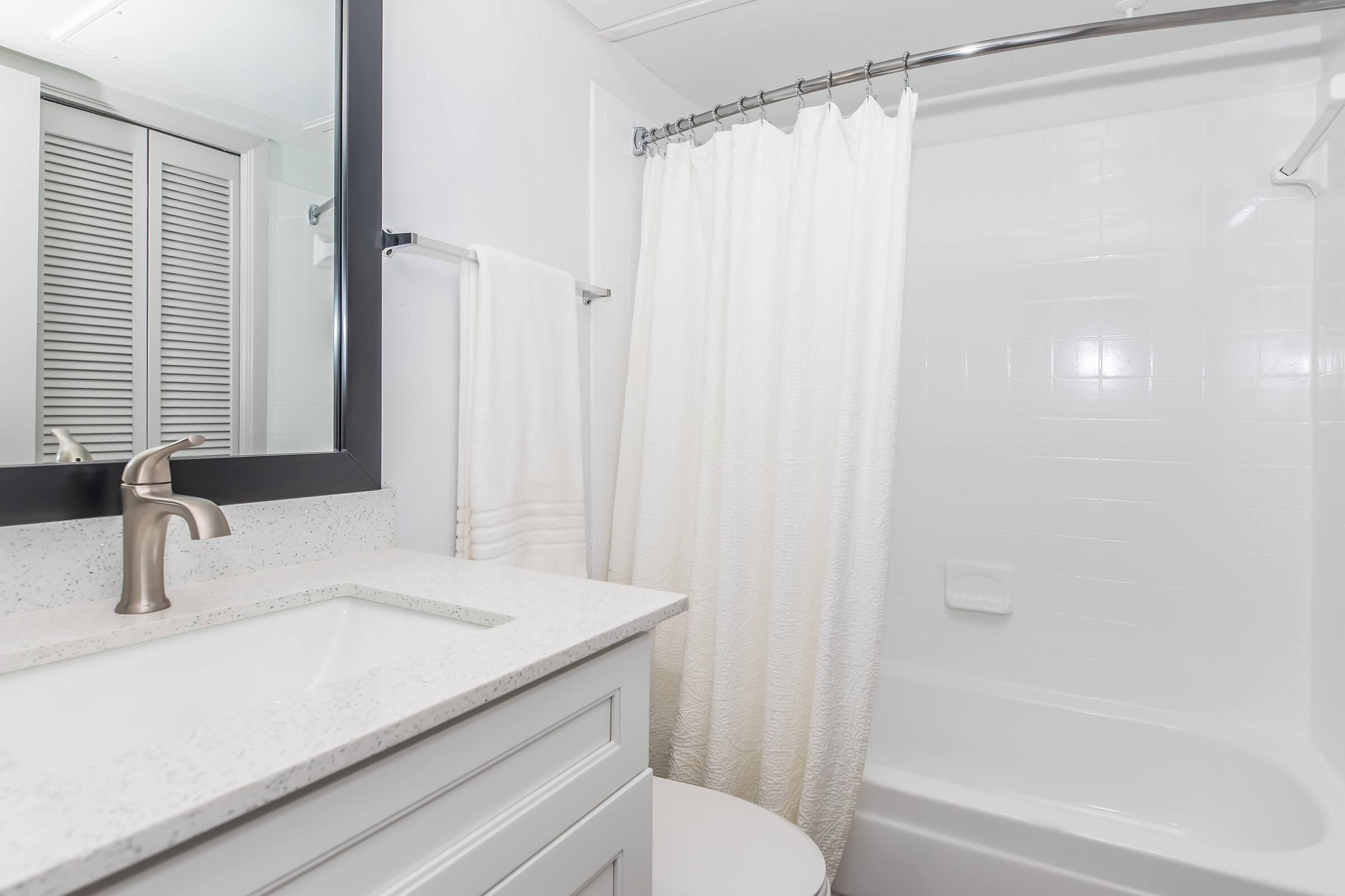A clean and modern bathroom featuring a white bathtub with a shower curtain, a light granite countertop with a sleek faucet, and a large mirror above the sink. The space includes a towel rack with a white towel and light-colored cabinet beneath the sink.