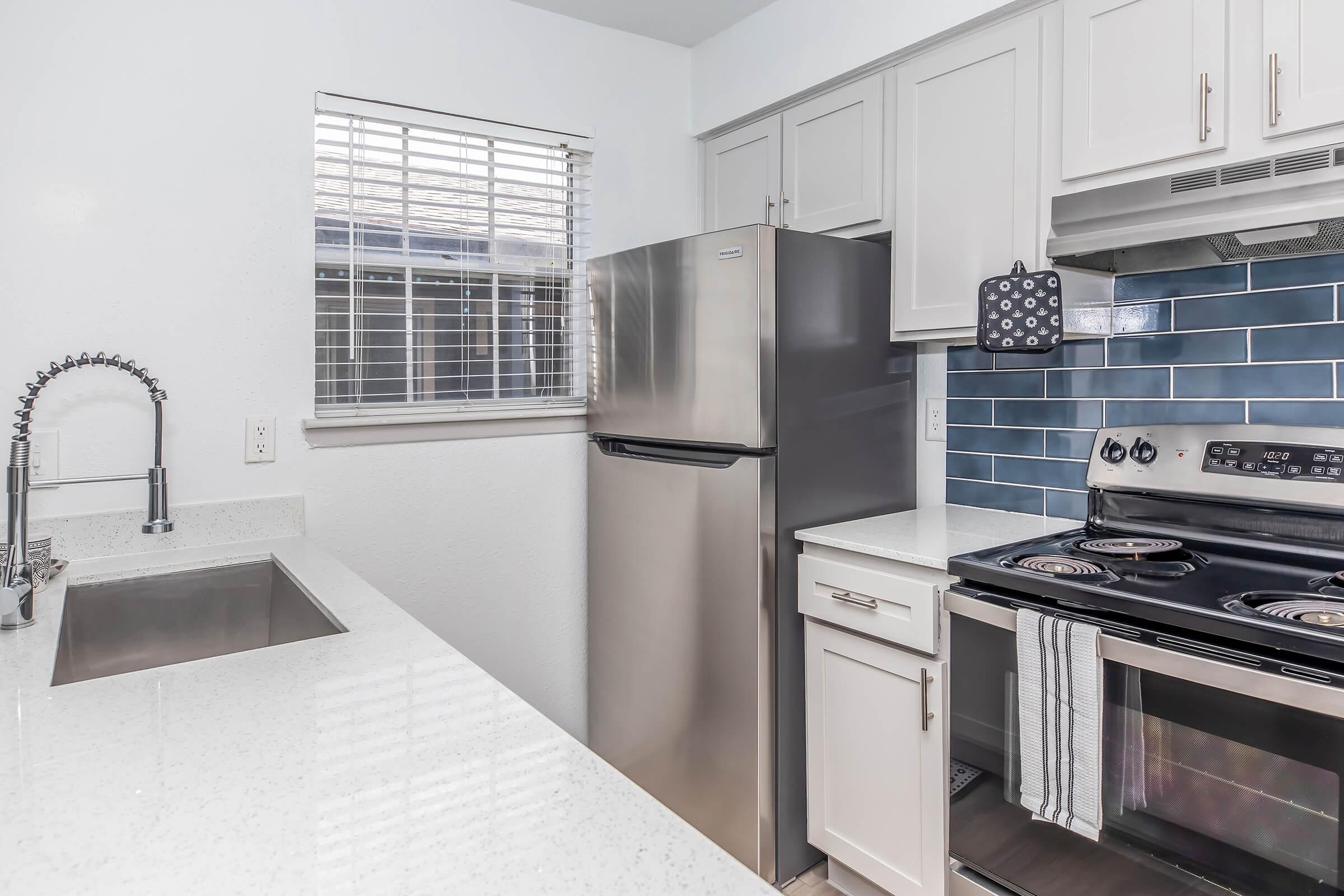 Modern kitchen featuring stainless steel refrigerator, stove, and a stainless steel sink with a sleek faucet. The cabinetry is light-colored, complemented by blue tiled backsplash. A window provides natural light, and the countertop is made of white quartz.