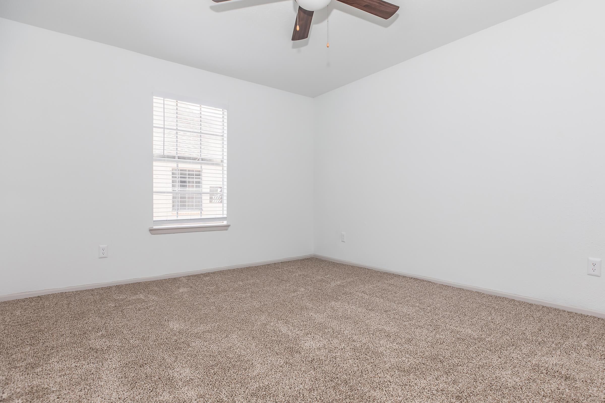 A sparsely furnished room with light beige carpet and white walls. A ceiling fan hangs from the ceiling, and there is a window with blinds allowing natural light to enter. The room appears clean and simple, suitable for various uses.