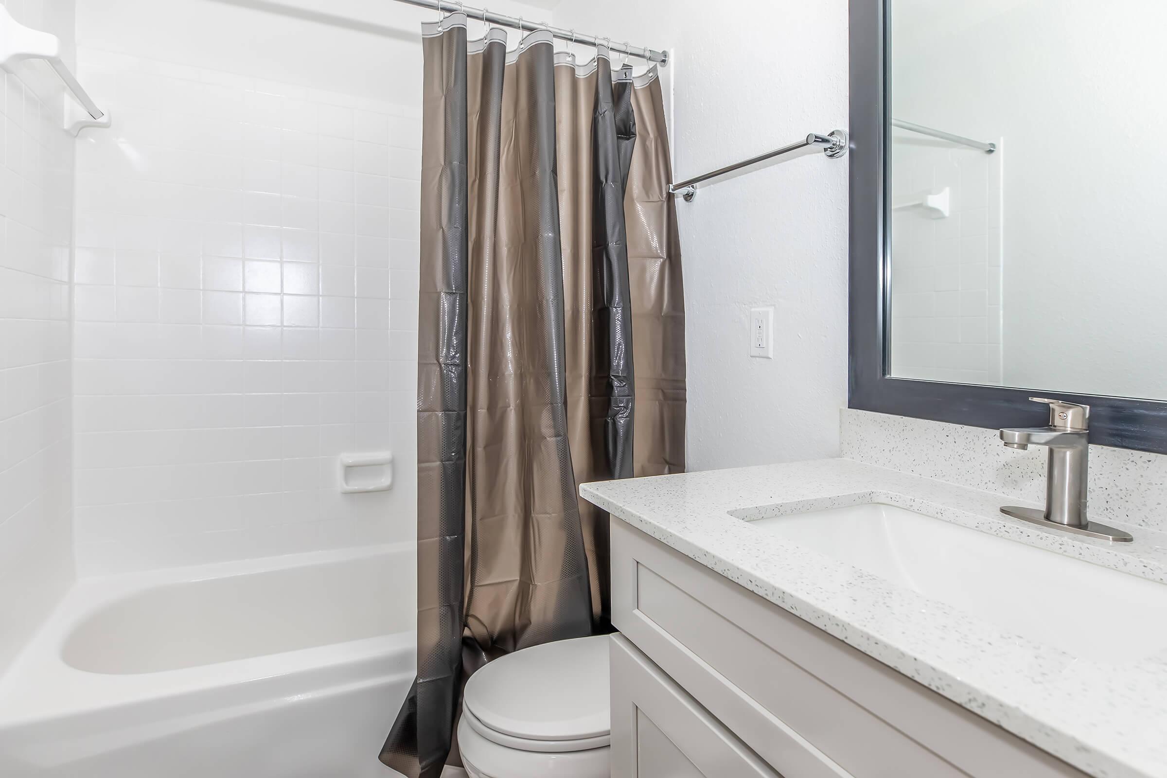 A clean, modern bathroom featuring a white tub with a shower, a dark brown curtain, a light gray vanity with a sink, a mirror above the sink, and a toilet. The walls are painted white, and the overall look is bright and spacious.