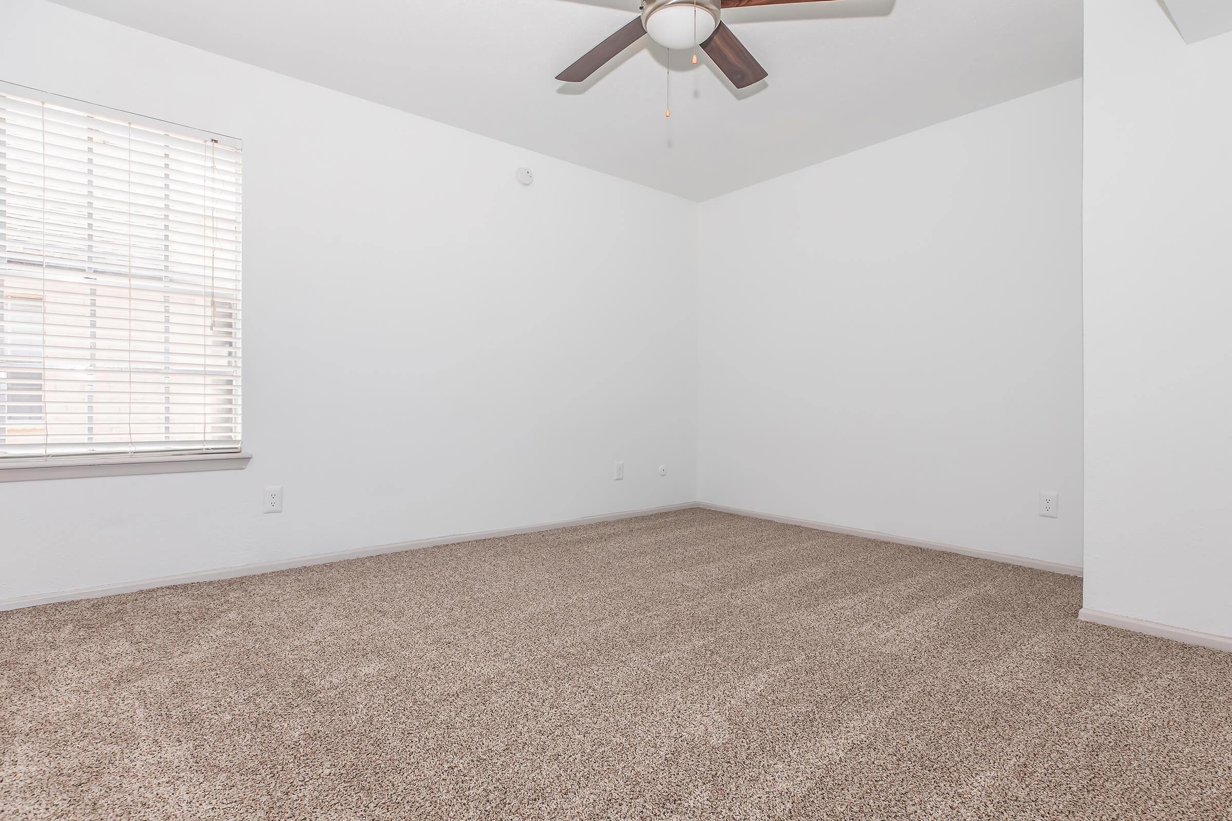 A spacious, empty room with beige carpet and a ceiling fan. The walls are white, and there is a window with blinds on one side, allowing natural light into the space. The floor is clean and uncluttered, suggesting potential for various uses or furnishings.