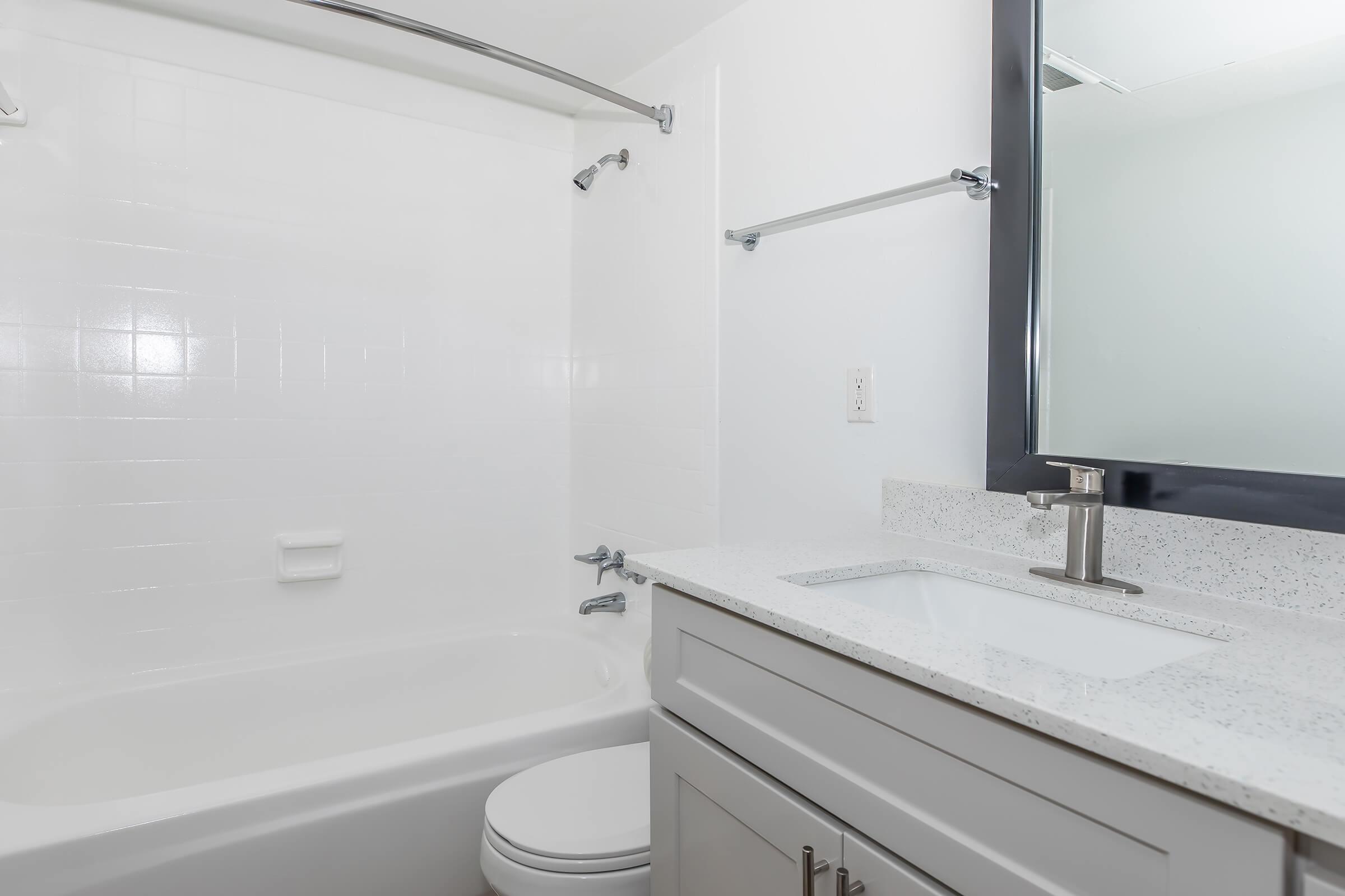 A clean and modern bathroom featuring a white bathtub with a showerhead, a sleek granite countertop with a sink, a large mirror above the sink, and minimalist storage cabinets. The walls are bright and tiled, enhancing the spacious feel of the area.