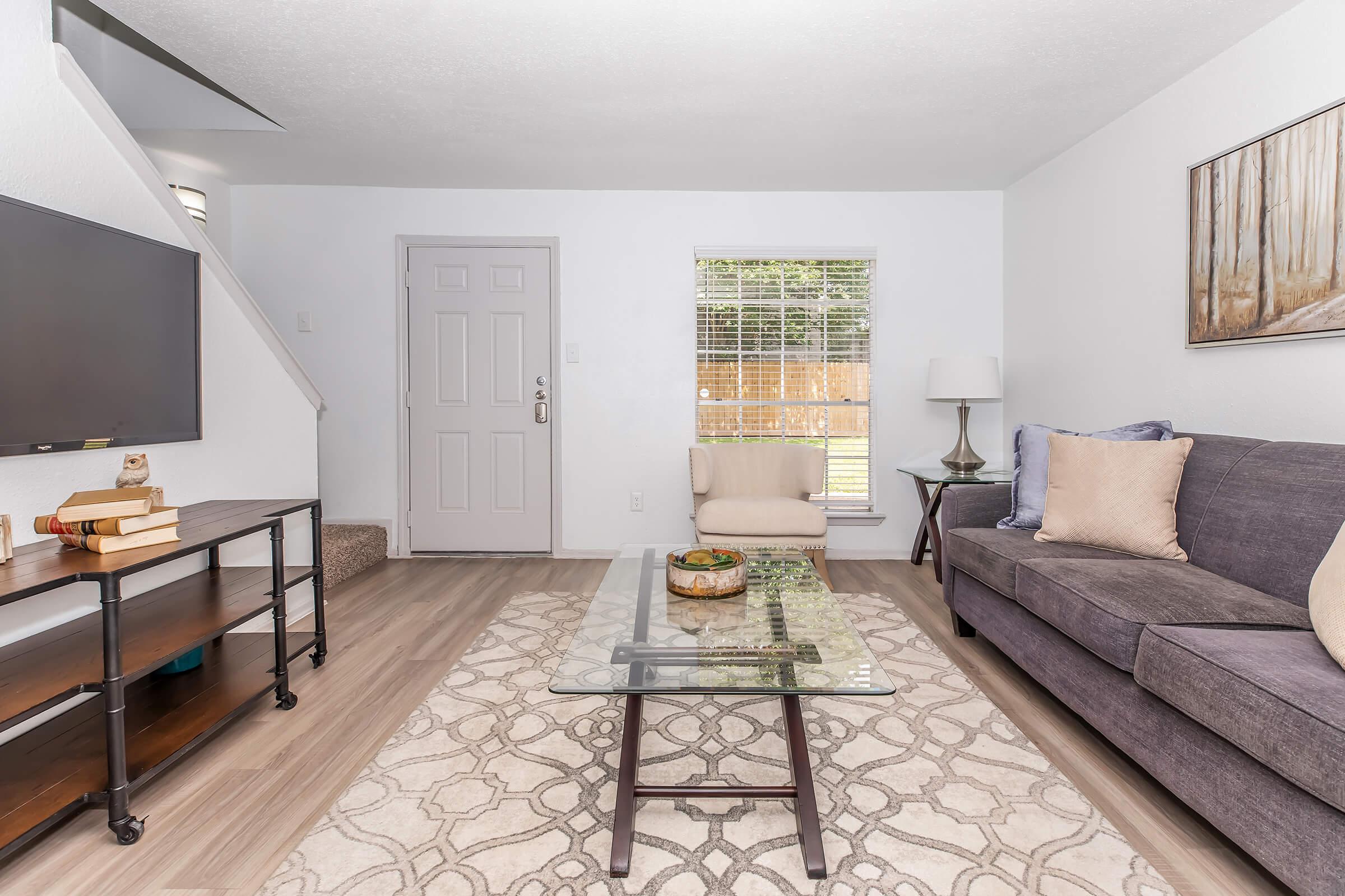 A modern living room featuring a gray sofa, a glass coffee table with a decorative bowl, and a flat-screen TV on a black metal stand. There is a large window with white blinds letting in natural light, and a small lamp beside the sofa. A neutral-colored area rug adds warmth to the space.