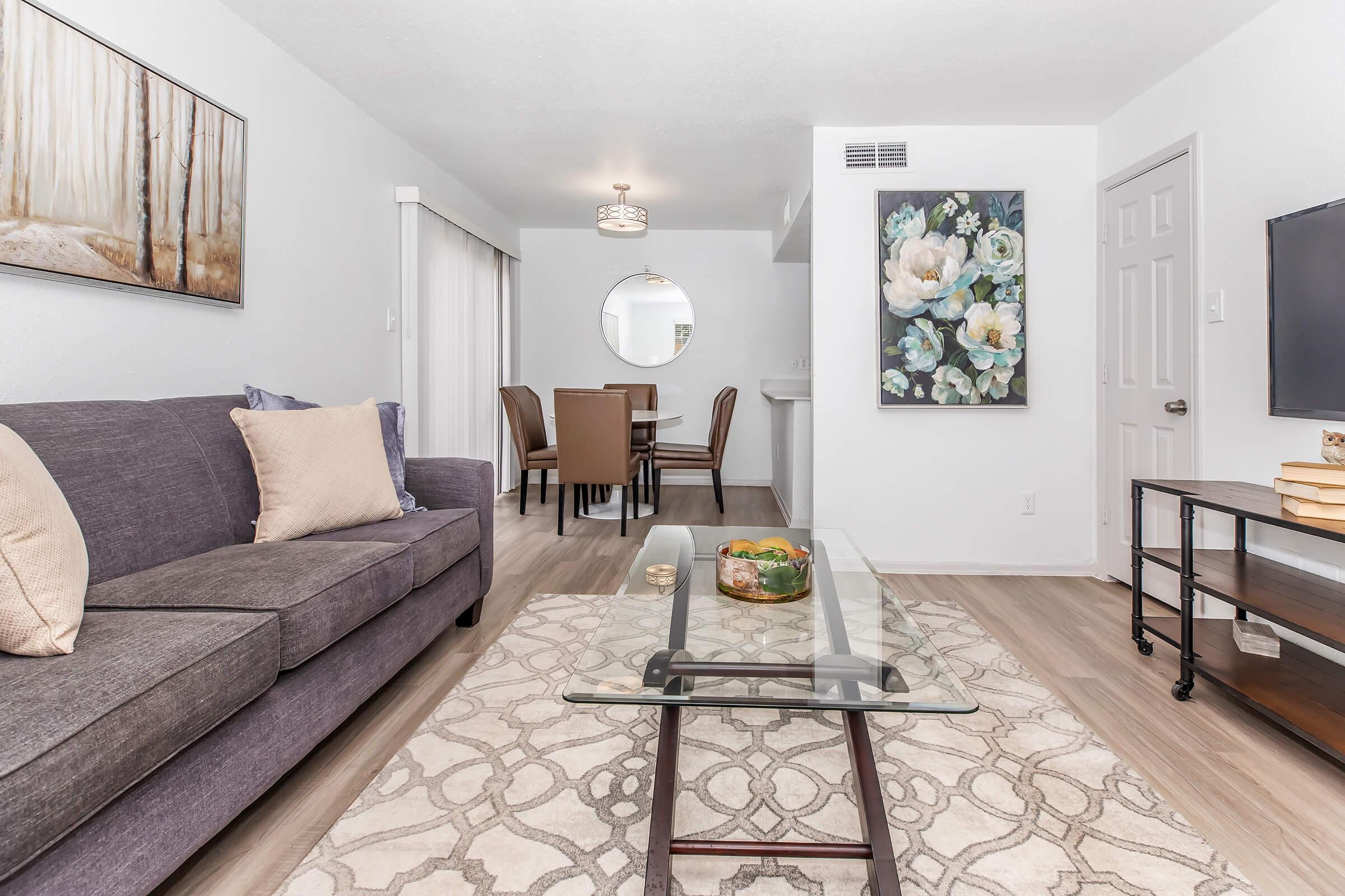 Modern living room featuring a gray couch with a beige pillow, a glass coffee table, and a decorative bowl. A large painting of flowers hangs on the wall, along with a round mirror. In the background, there is a dining area with four brown chairs and a light fixture above, complemented by light wood flooring.