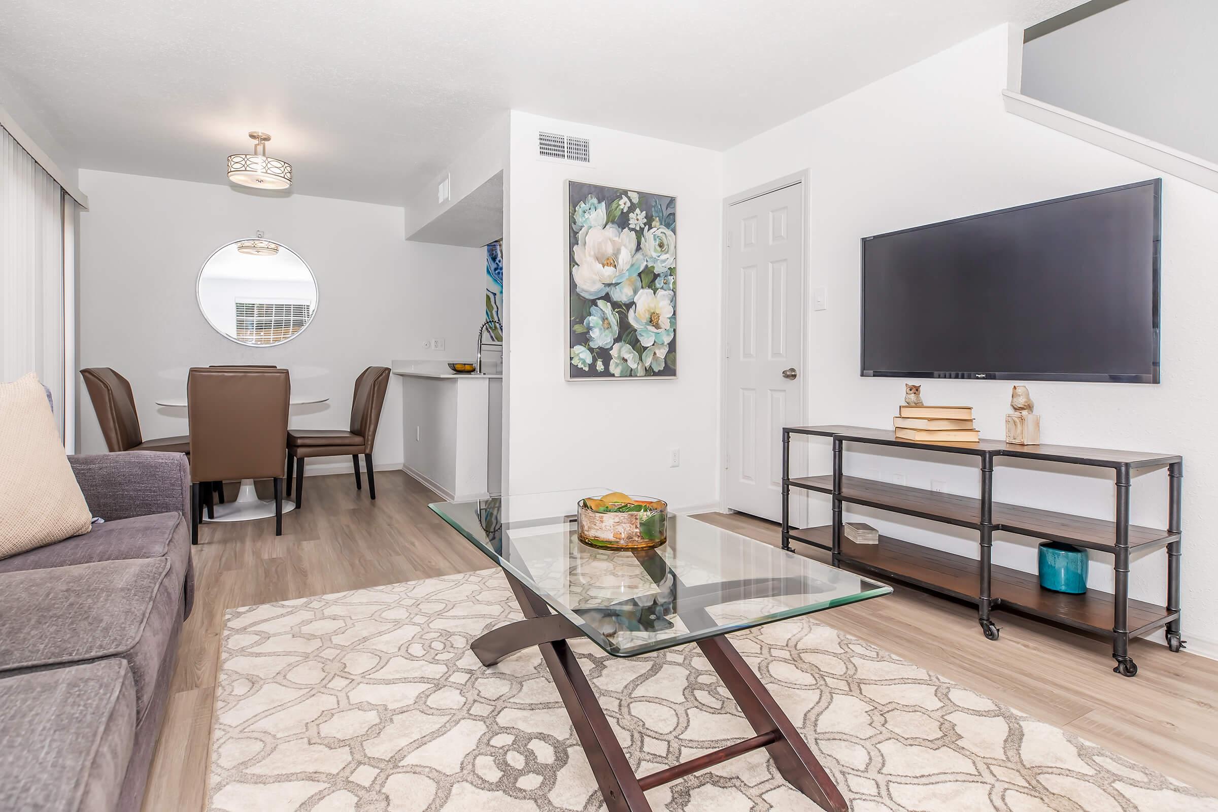 Modern living room featuring a gray sofa, a glass coffee table, and a flat-screen TV on a black stand. A round mirror and a floral artwork adorn the walls. In the background, a dining area with two chairs is visible, alongside a small kitchen bar. The space is bright and well-decorated with a cozy ambiance.