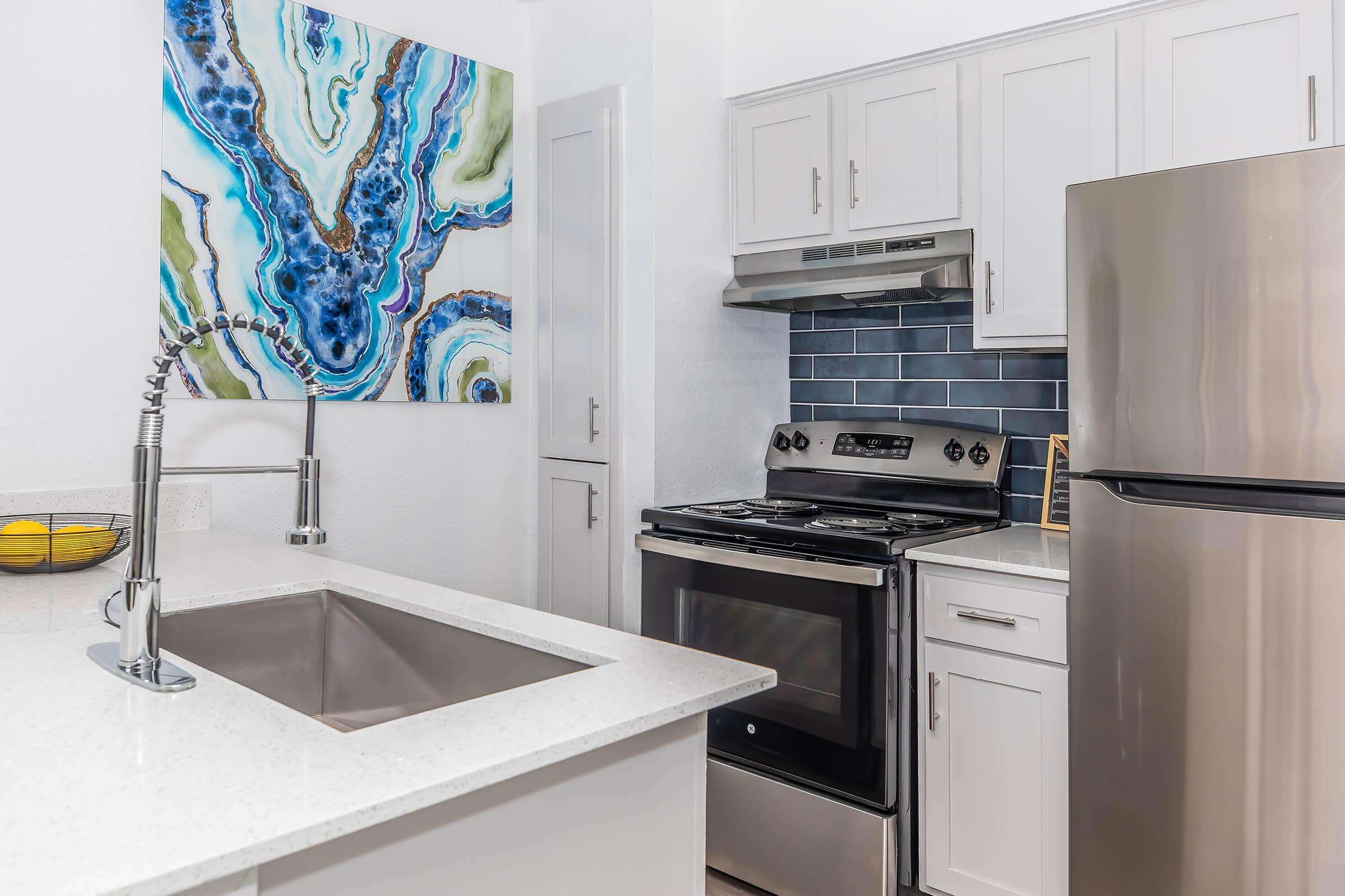Modern kitchen featuring stainless steel appliances, including a gas stove and refrigerator. There is a double basin sink with a sleek faucet. The cabinetry is light-colored, and a vivid abstract painting adorns the wall above the sink, adding a pop of color to the space.