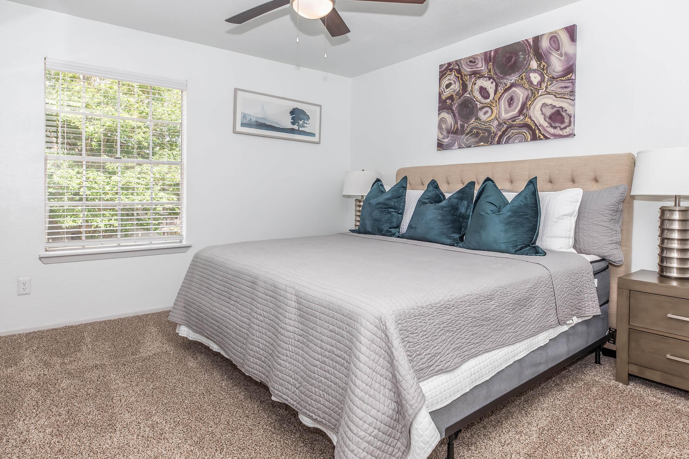 A cozy bedroom featuring a neatly made queen-sized bed with a light gray quilt and decorative teal pillows. There are two bedside lamps on nightstands, and a piece of abstract wall art above the bed. A window with white blinds lets in natural light, illuminating the soft carpeted floor.